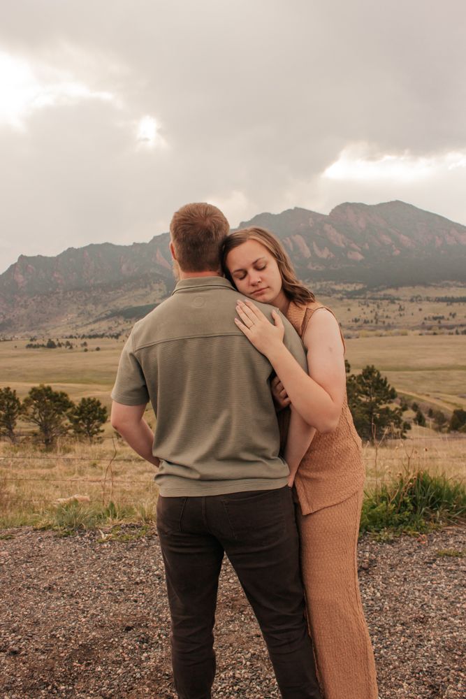 A couple embraces against a scenic mountain landscape backdrop with rolling hills and cloudy skies at sunset.