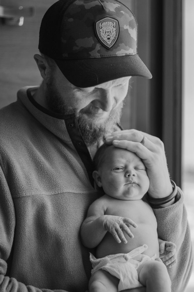 Black and white photo series showing tender moments between a parent wearing a baseball cap holding a sleeping newborn baby.