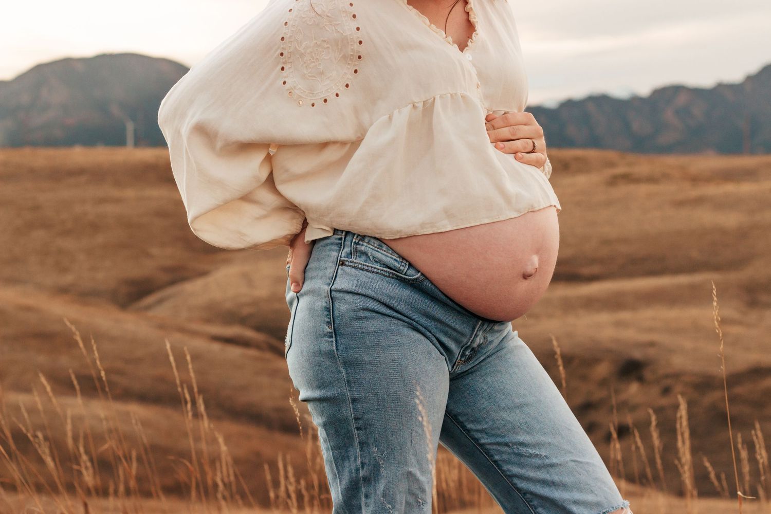 Pregnant woman in white blouse and jeans cradling  baby bump in golden field with mountains.Motherhood + Family Photographer Westminster,Co
