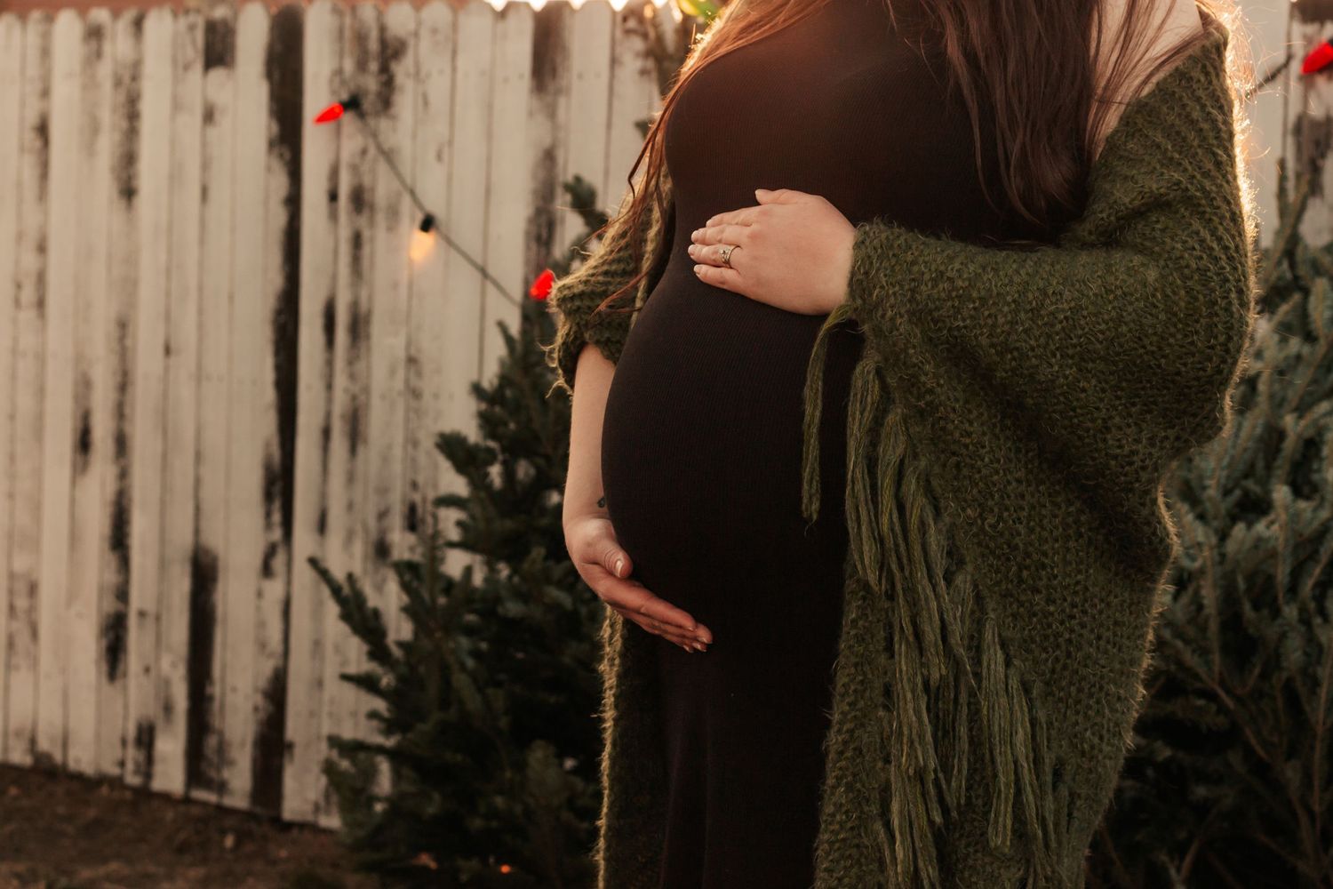 Pregnant woman in green shawl cradling baby bump outdoors near Christmas trees |Family photographer in  Westminster CO
