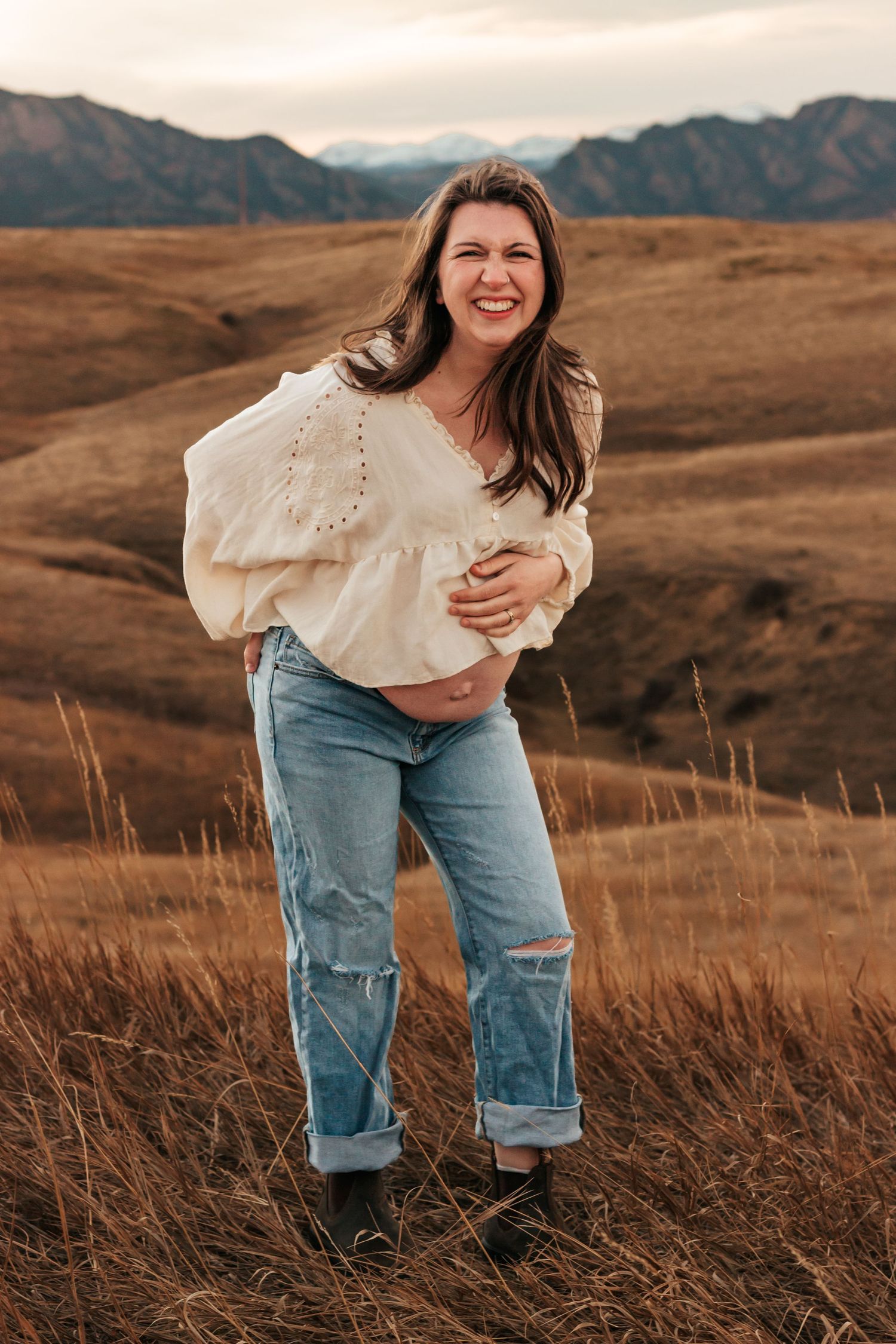 Smiling pregnant woman in white blouse and ripped jeans holding baby bump | Motherhood + Family Photographer Westminster, CO