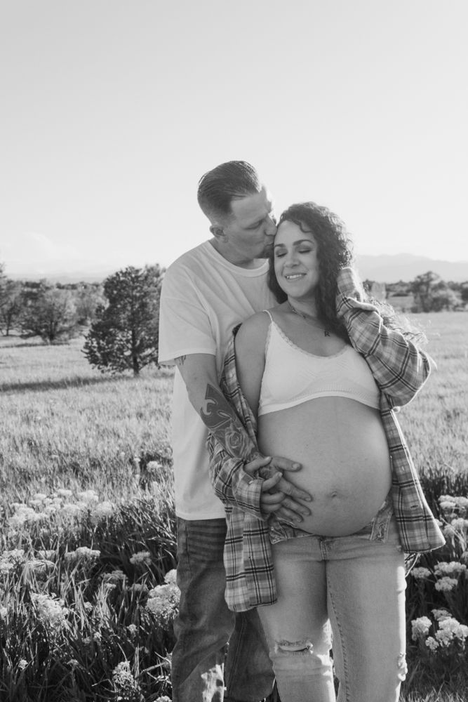 A black and white maternity photo of an expectant couple embracing in a sunny meadow with mountains in the background.