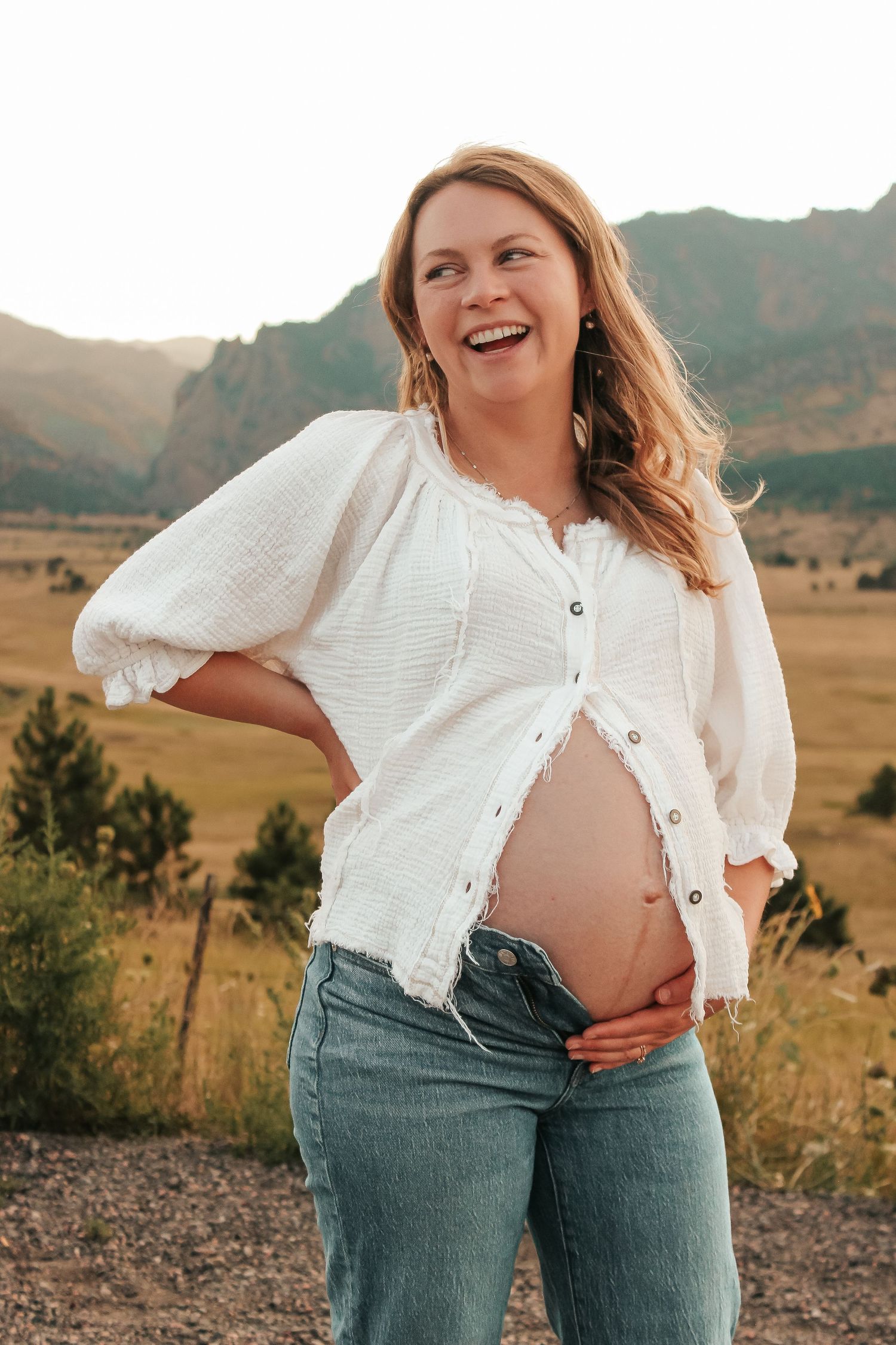 A joyful maternity photo showing a pregnant woman in a white blouse and jeans against a scenic mountain backdrop.