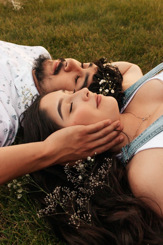 A couple lying in grass with delicate white flowers scattered in their dark hair, sharing an intimate moment together.