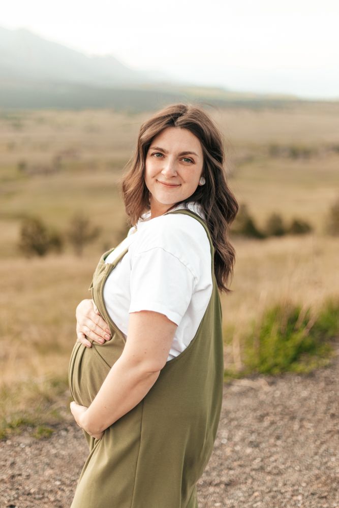 Pregnant woman in green overalls and white tee cradling her baby bump |Motherhood + Maternity Photographer in Westminster, CO