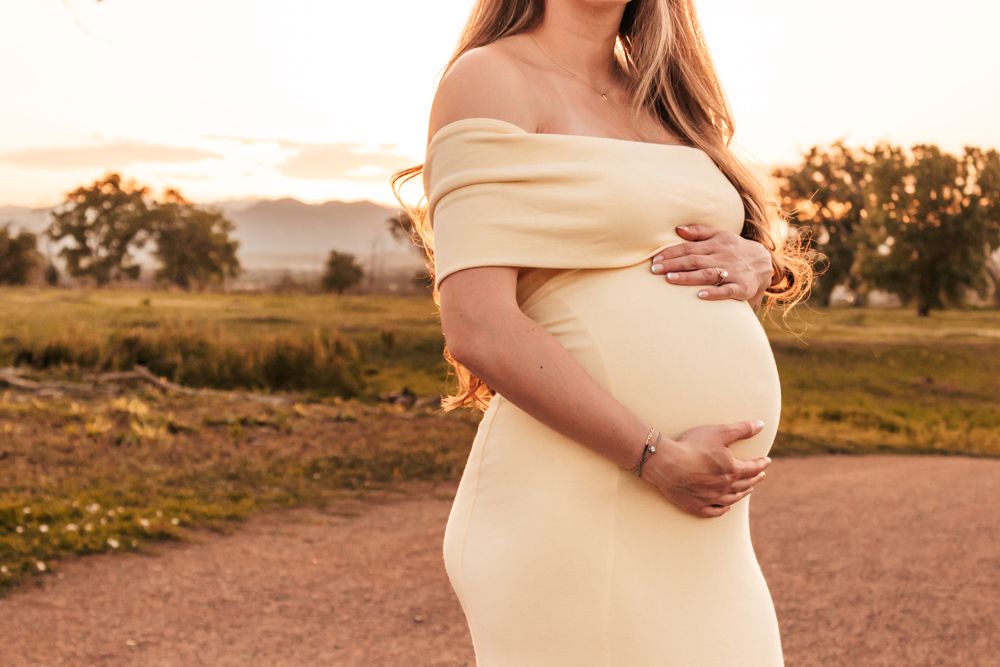 Pregnant woman in cream off-shoulder dress cradling baby bump at golden hour | Motherhood + Family Photographer Westminster, CO
