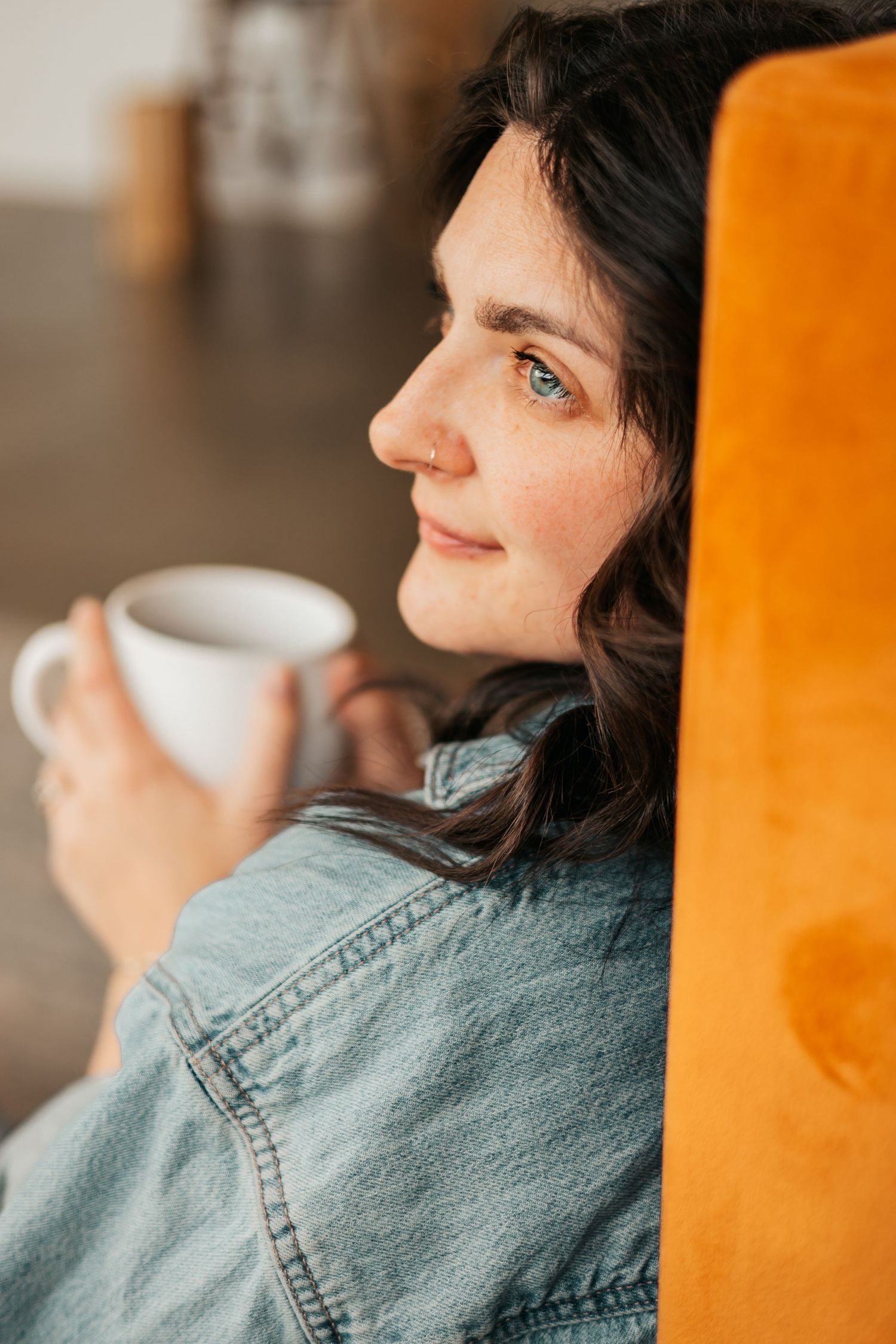 Young woman with blue eyes holding a white coffee cup, leaning against an orange sofa in a denim jacket.