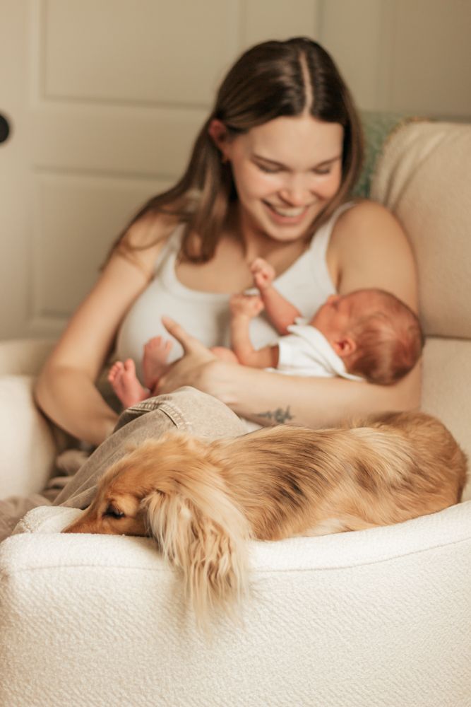 A mother holds her newborn baby while a golden retriever dog rests peacefully beside them on a white bed.