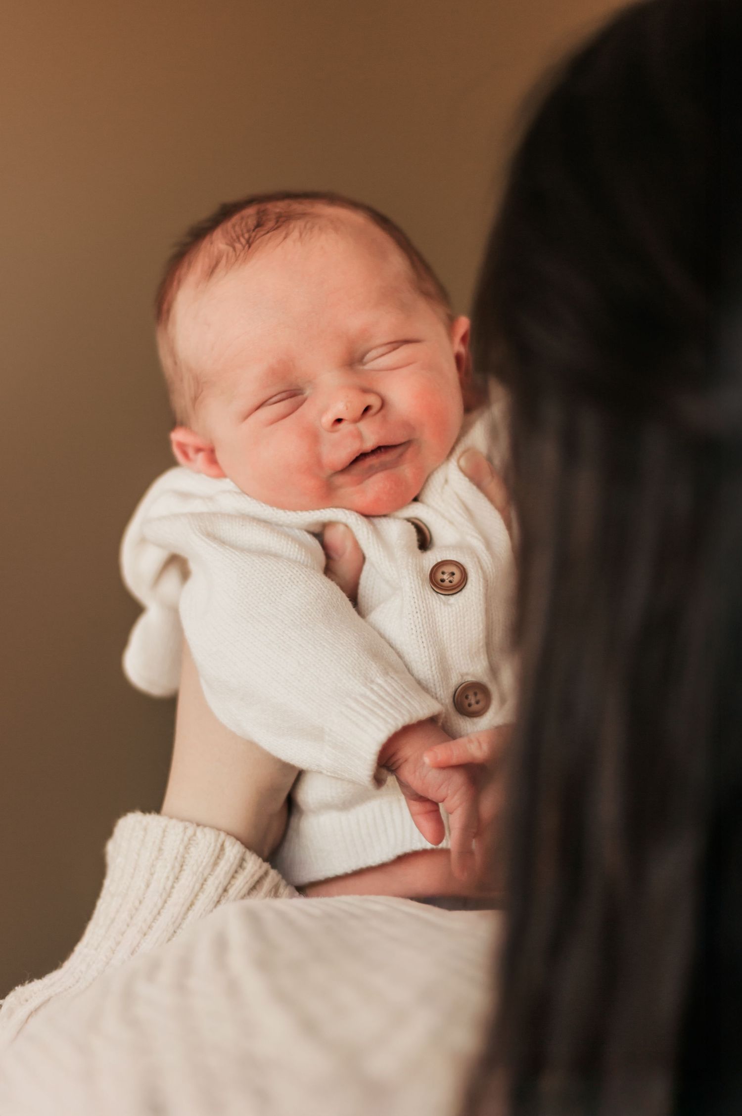 A smiling newborn baby wearing a white knit cardigan is held up by someone in a cream colored sweater.Family photographer in  Westminster CO