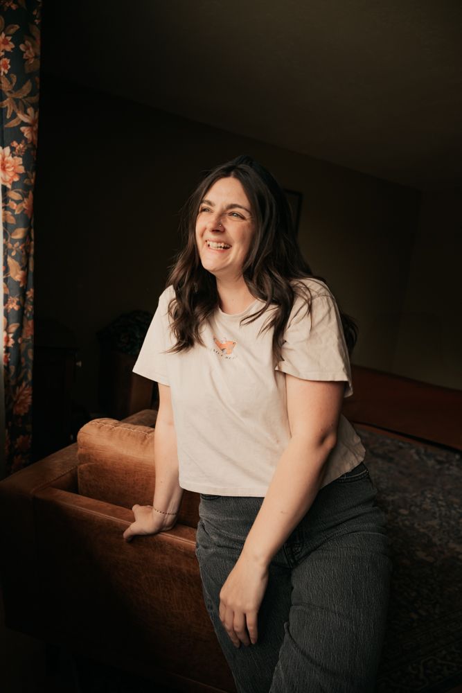 Person in white shirt and dark pants sits on couch in dimly lit room with joyful expression.