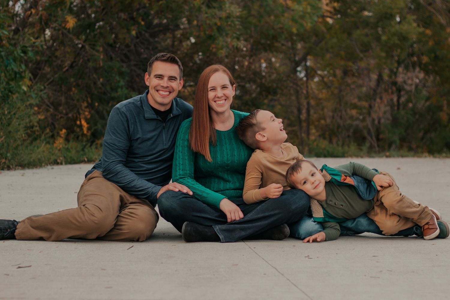 Happy family of four sitting outdoors on pavement, smiling and laughing together.|Motherhood + Maternity Photographer in Westminster, CO