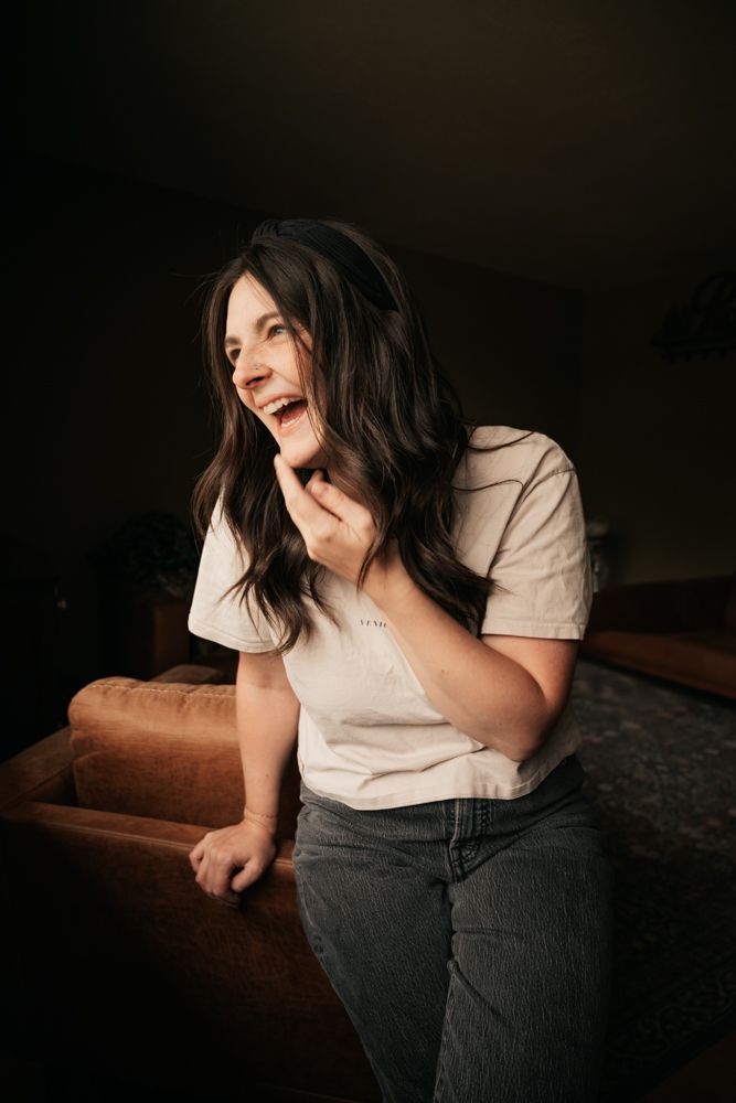 A laughing brunette woman in a white t-shirt and jeans leans against a brown sofa| Motherhood + Maternity Photographer in Westminster, CO
