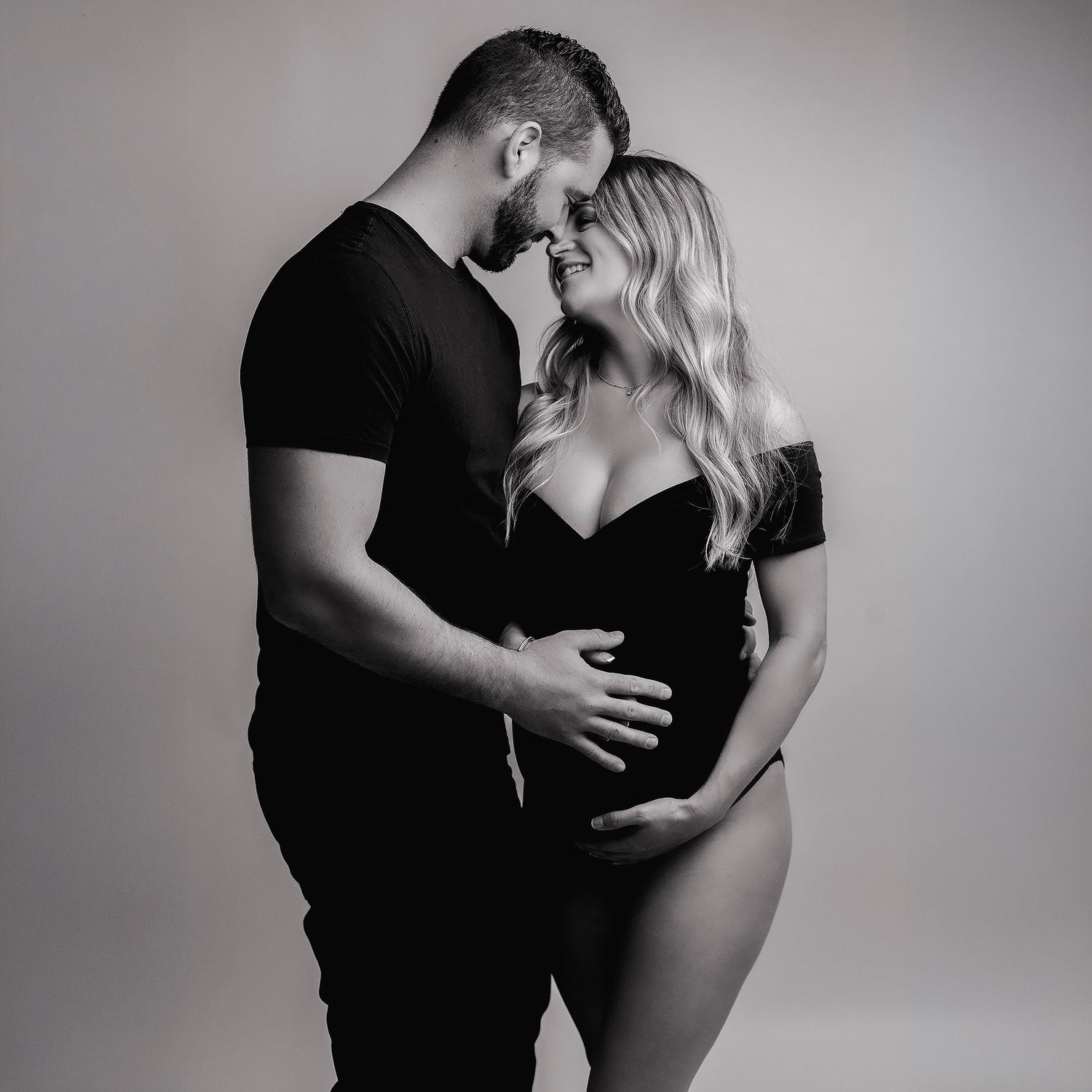 Black and white maternity photo of a couple in black outfits, tenderly embracing and cradling a baby bump.