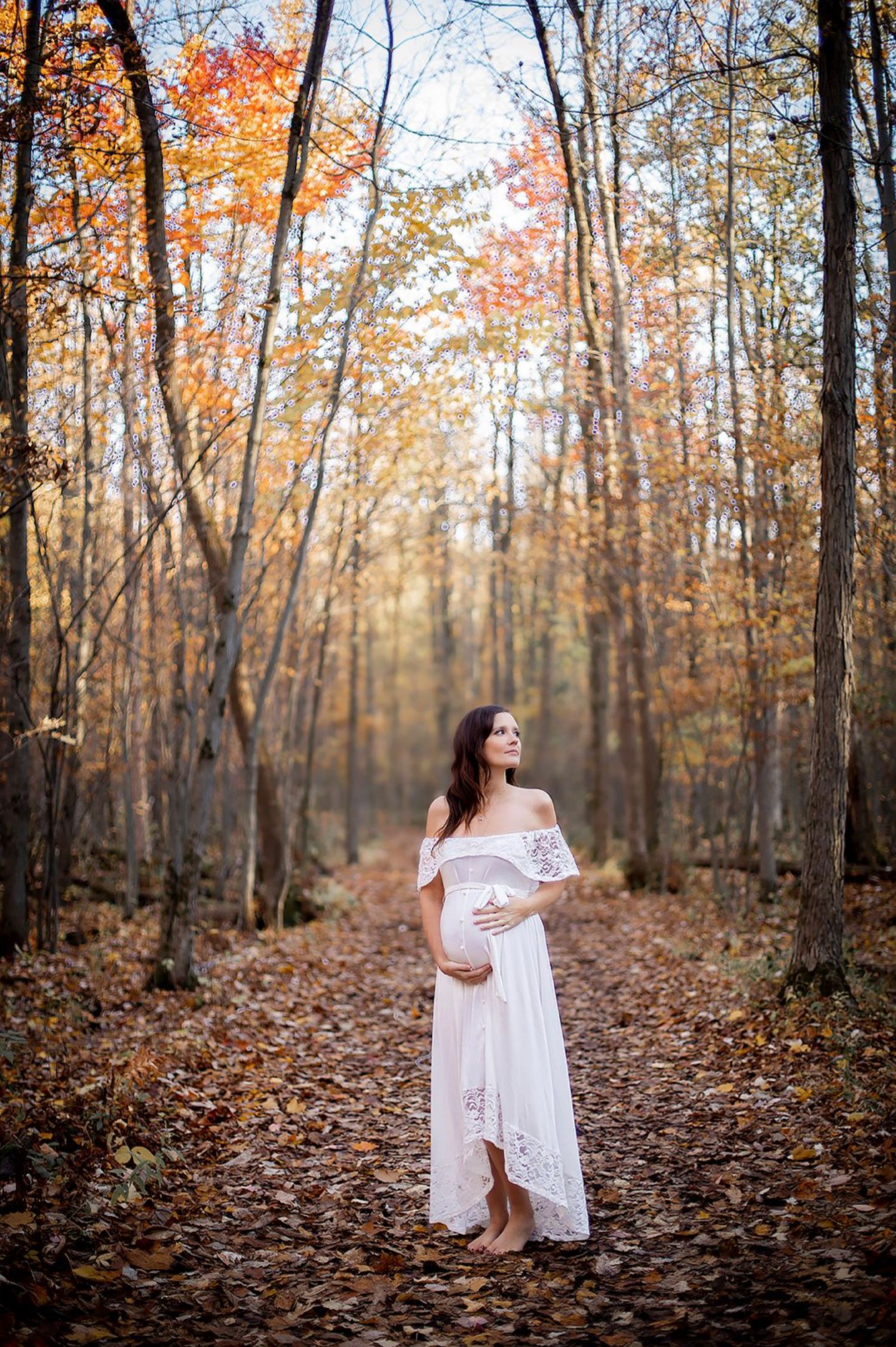 Pregnant woman in white lace dress standing on autumn forest path surrounded by golden fall foliage.