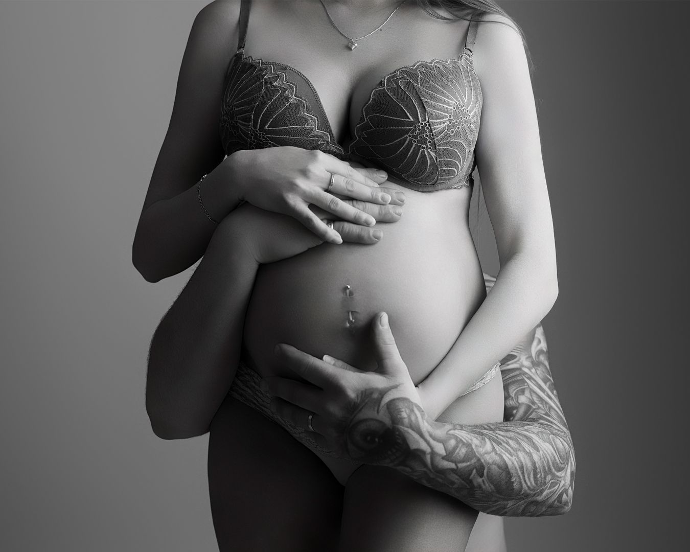 Black and white maternity photo of pregnant woman in lace bra cradling baby bump with tattooed arms.