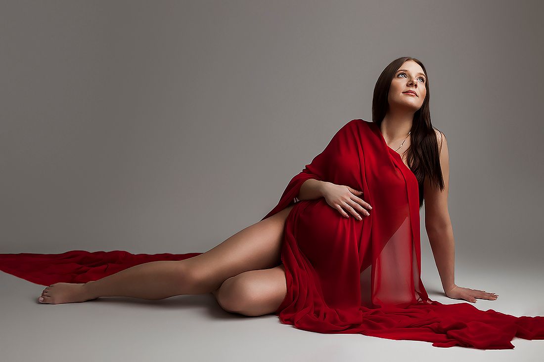 Pregnant woman in flowing red dress sitting gracefully on studio floor, maternity photography portrait.