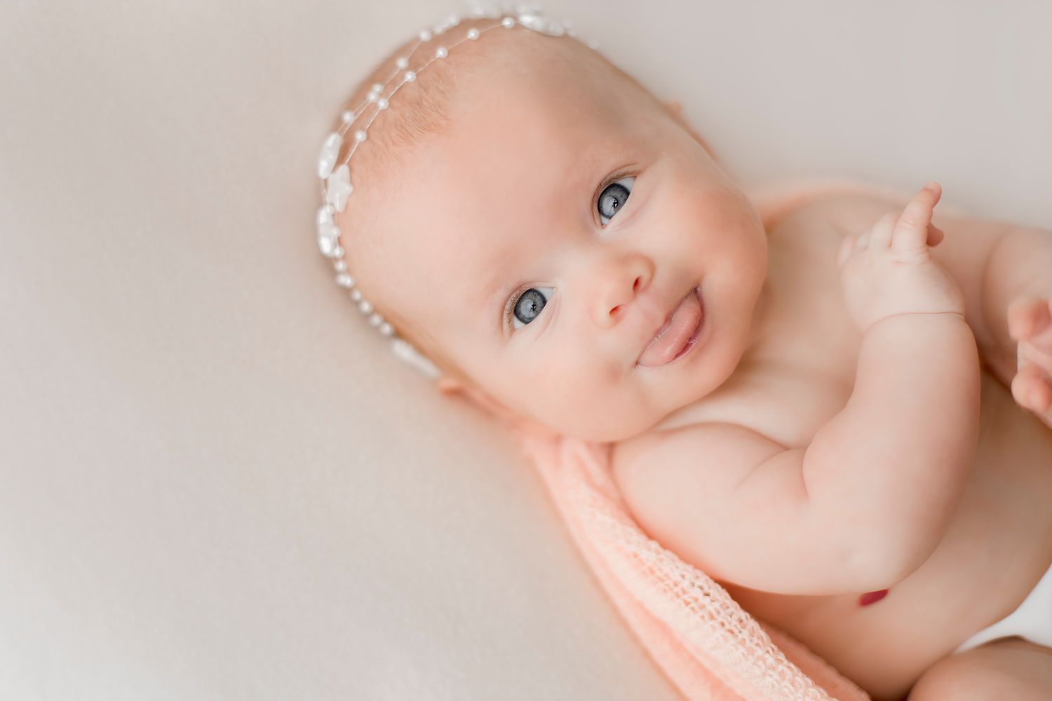 Smiling newborn baby with blue eyes wearing a pearl headband wrapped in a soft pink blanket.