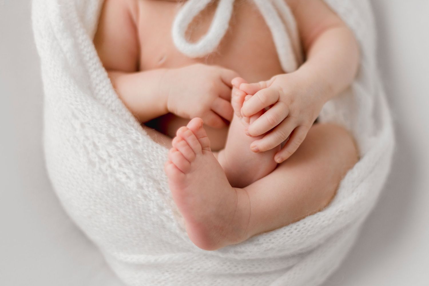 Close-up of a newborn baby's tiny feet and hands wrapped in a soft white blanket on a white background.