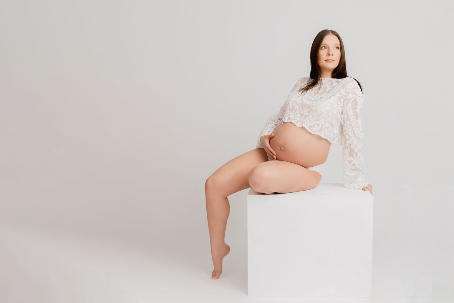 Pregnant woman in white lace top sitting on white cube, cradling baby bump in minimalist studio setting.
