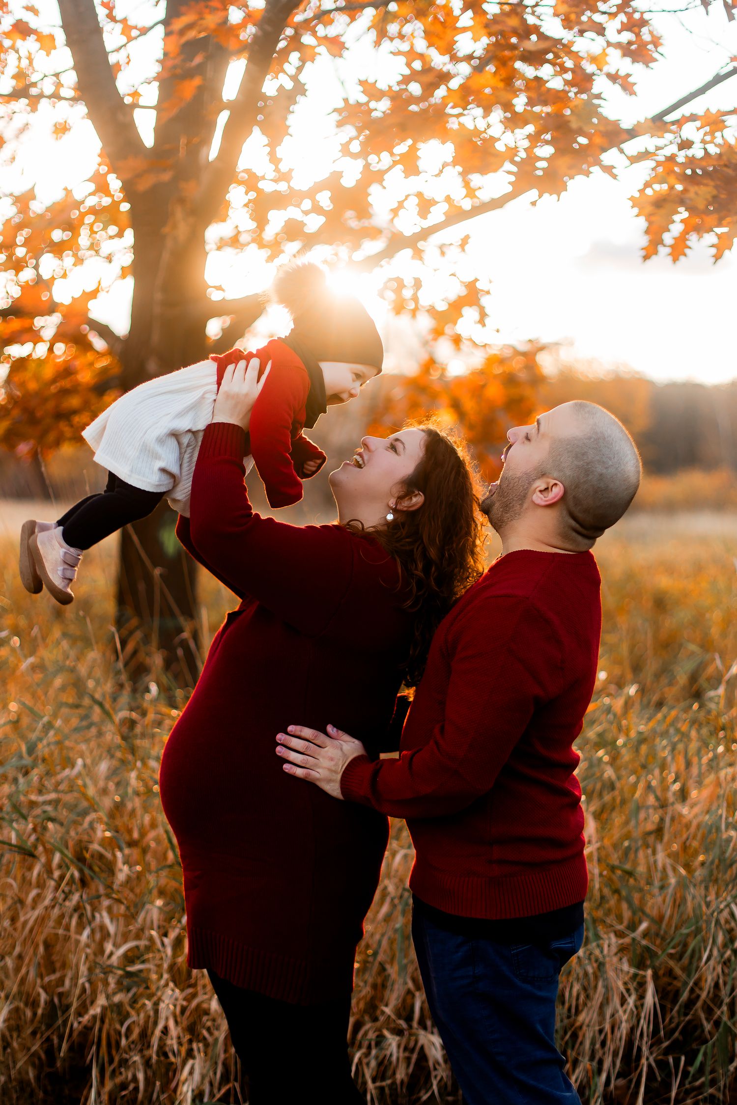 Family in red sweaters lifting toddler under autumn oak tree at golden sunset in a field.