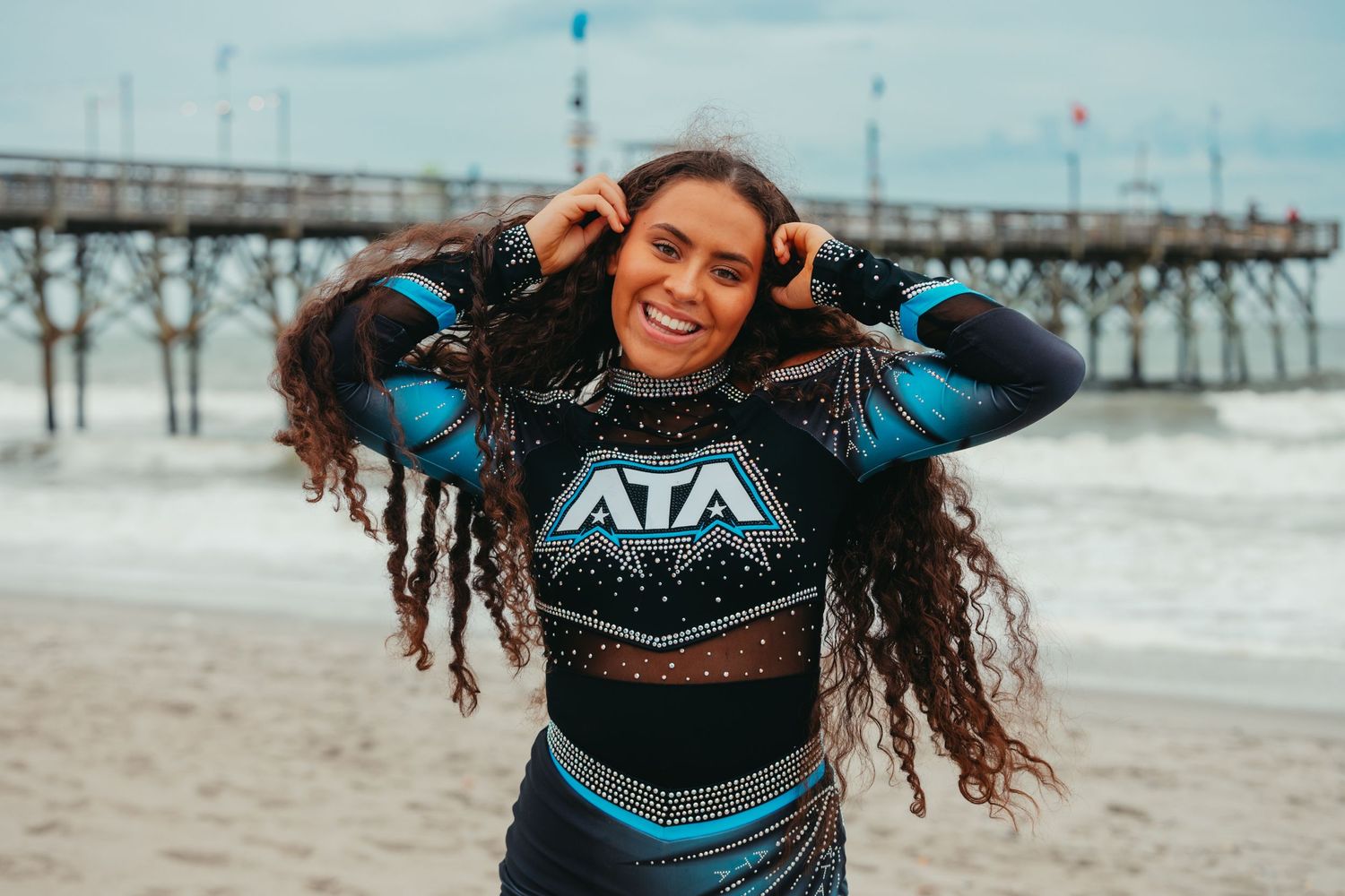 Smiling woman in ATA athletic top on a beach with a pier in the background.