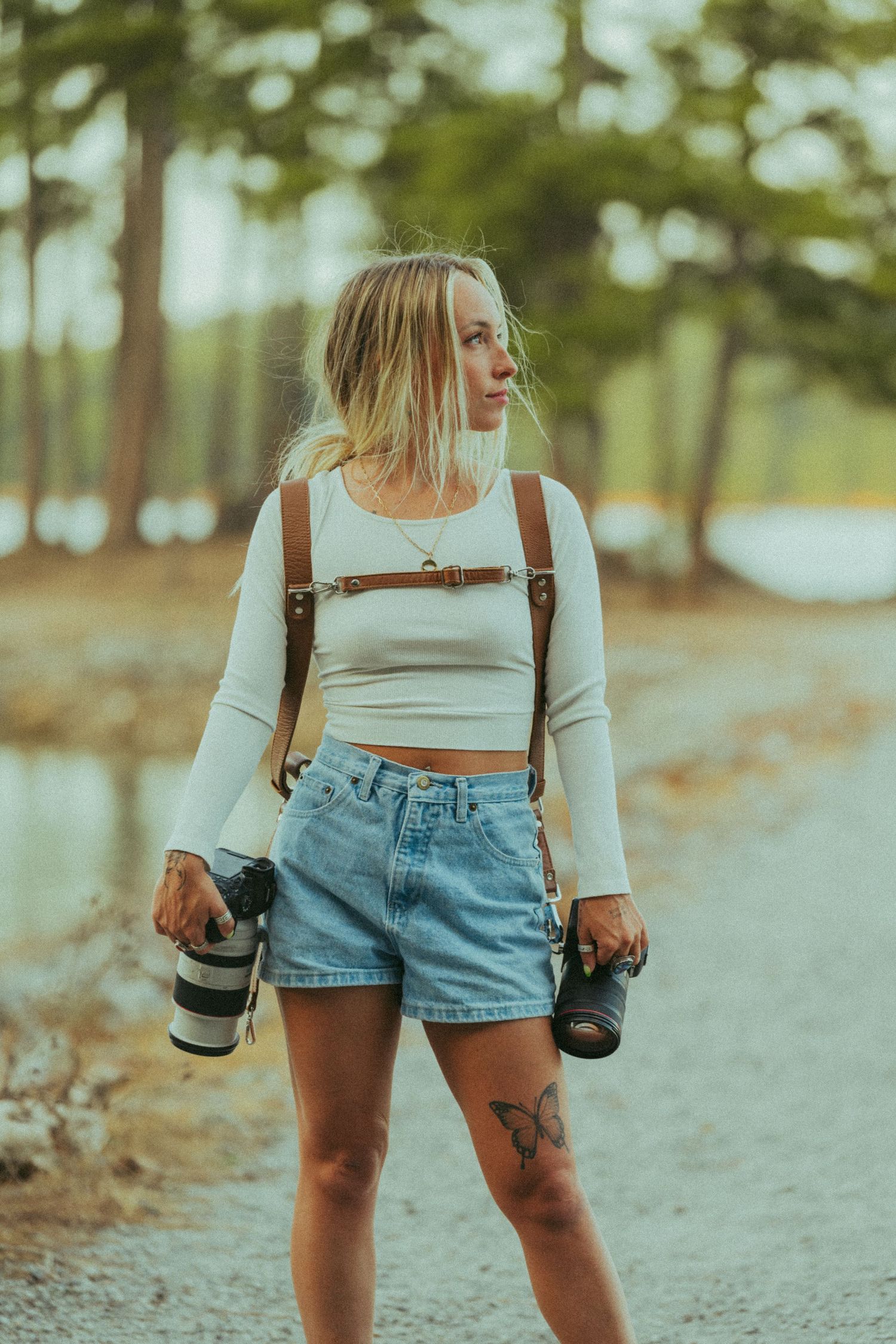 Blonde female photographer with leather backpack carrying two cameras outdoors.