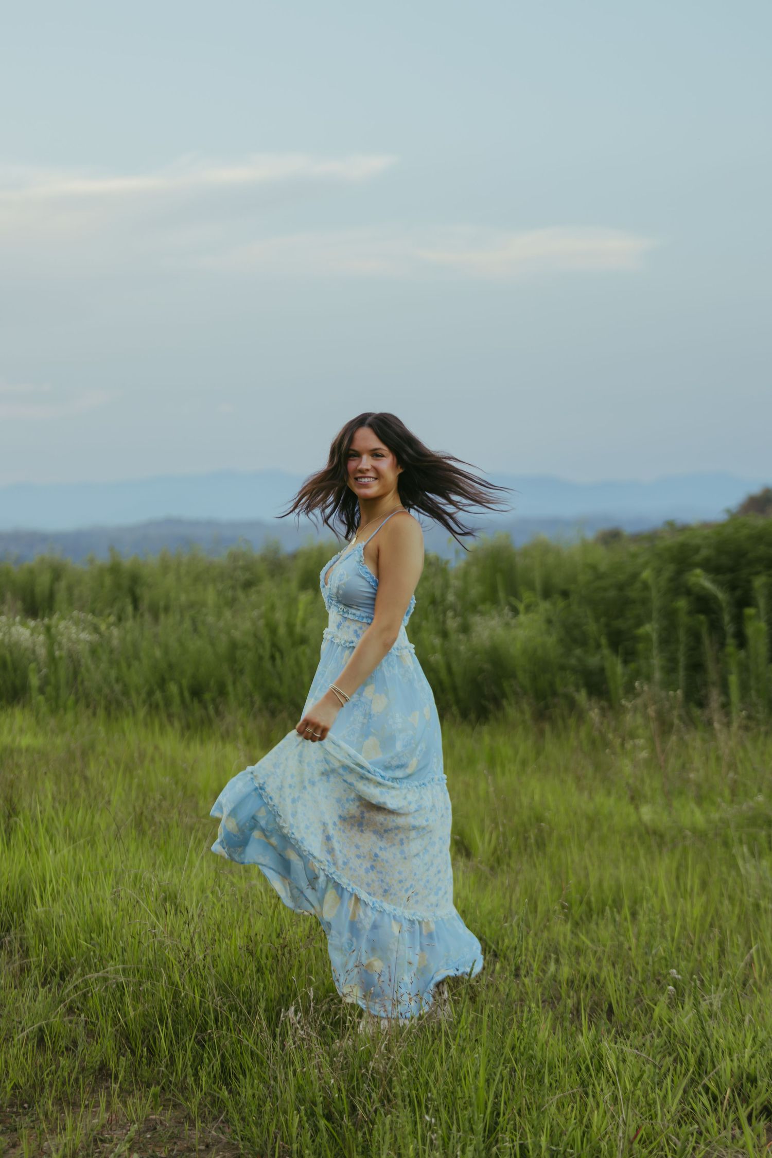 Young woman in flowing blue floral maxi dress twirling in green field at dusk.