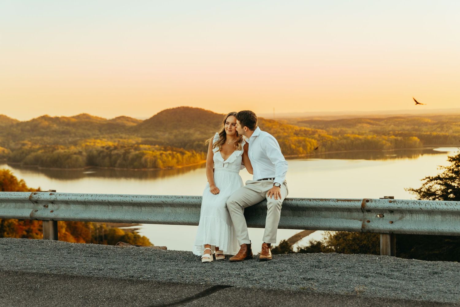 Couple in wedding attire sitting on a guardrail with a stunning sunset river valley backdrop.