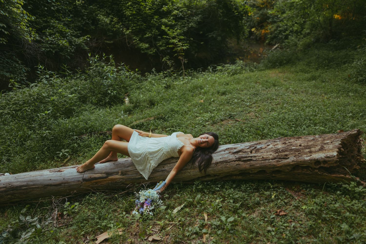 Young woman in white dress reclining on fallen log in lush green forest.