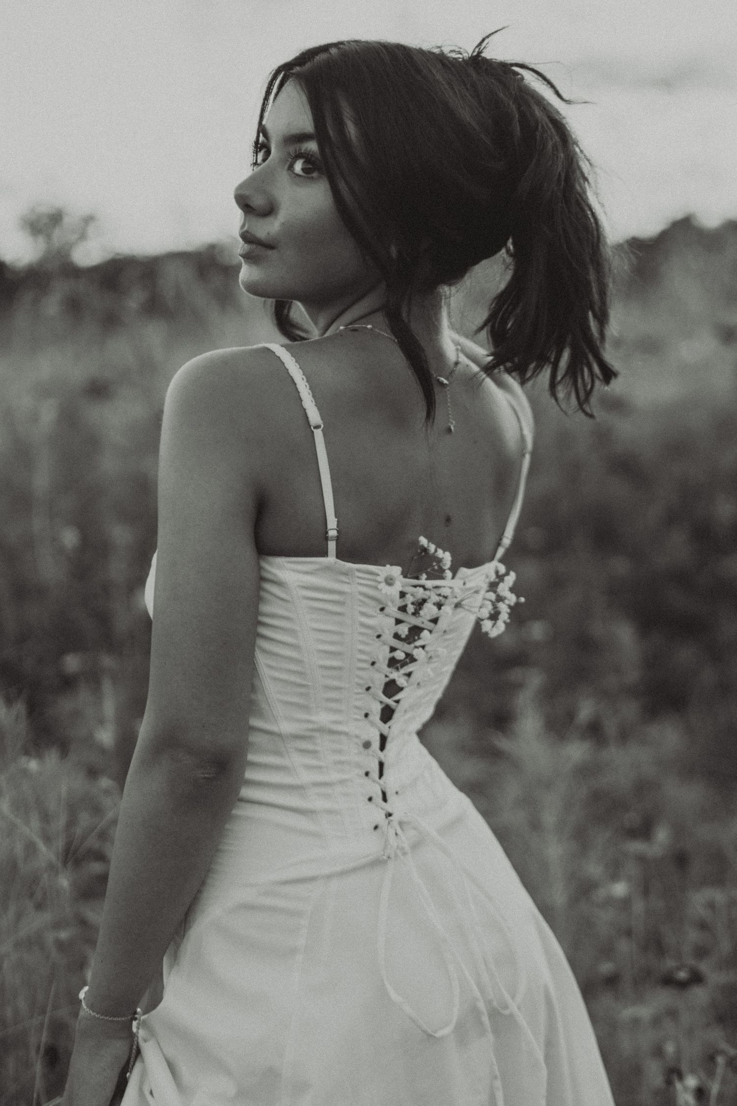 Black and white portrait of young woman in lace-up corset dress turning back in field.