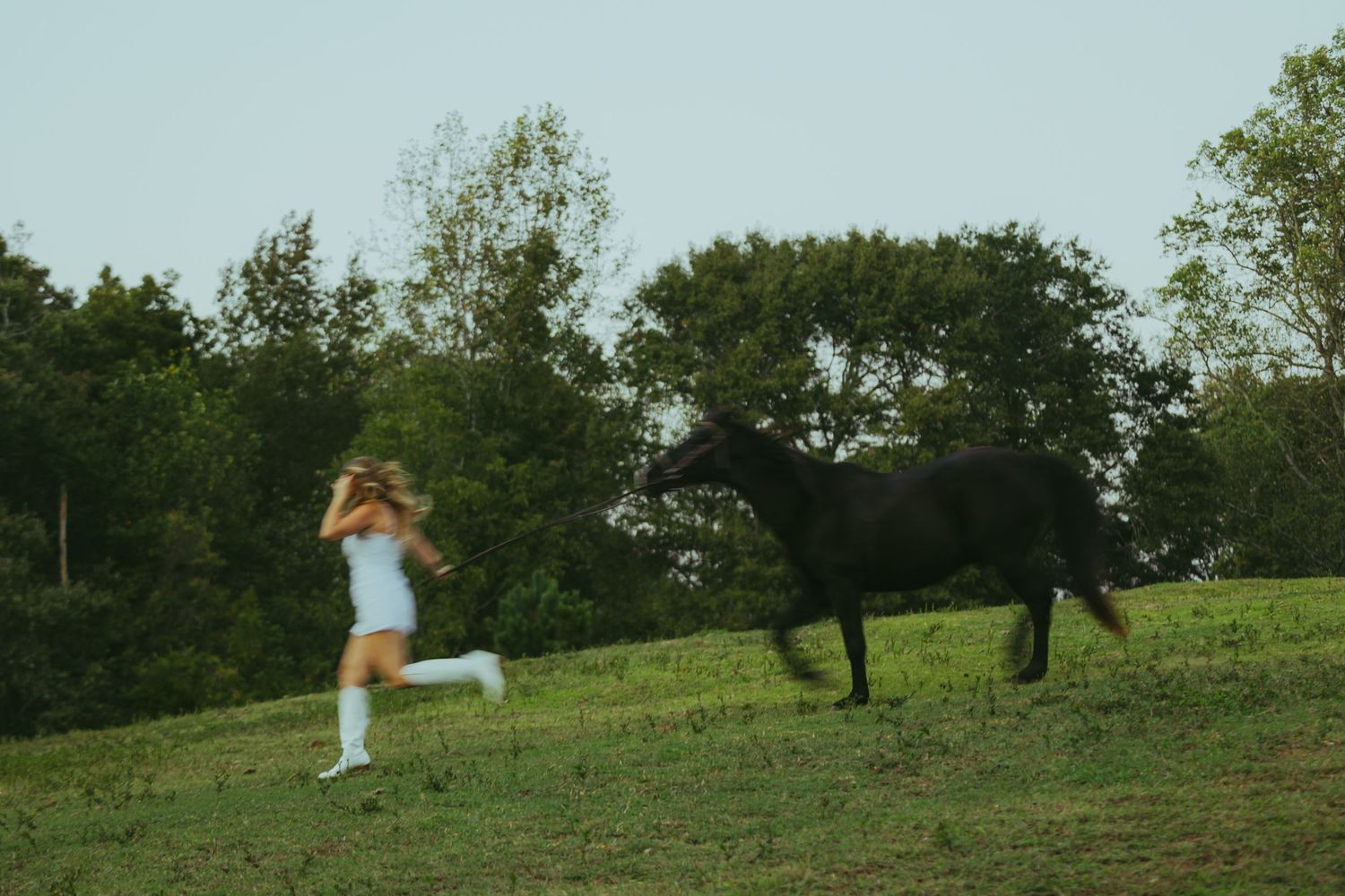 Young woman in white dress runs alongside a black horse on a lush green hillside.