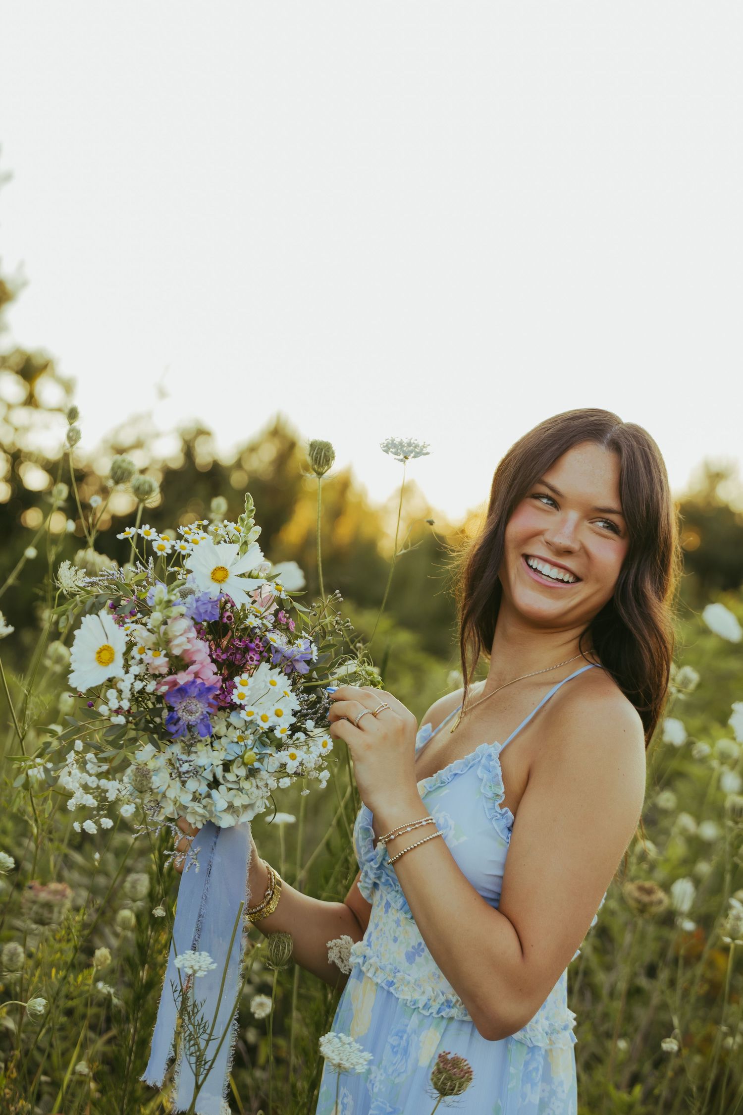 Smiling young woman holding wildflower bouquet in sunlit meadow wearing blue floral sundress.