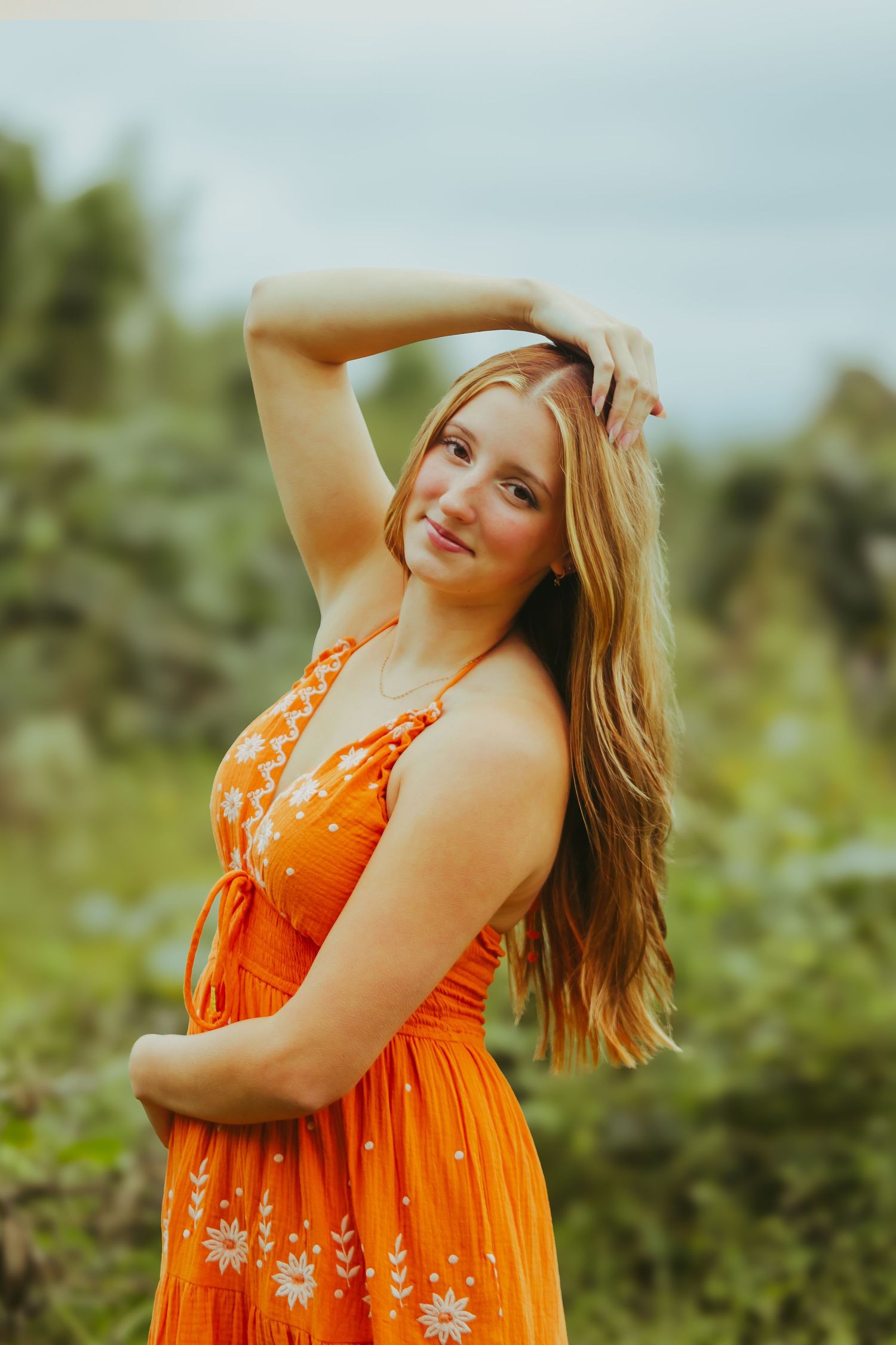 Young woman in orange floral sundress poses outdoors with hand in hair against green foliage.