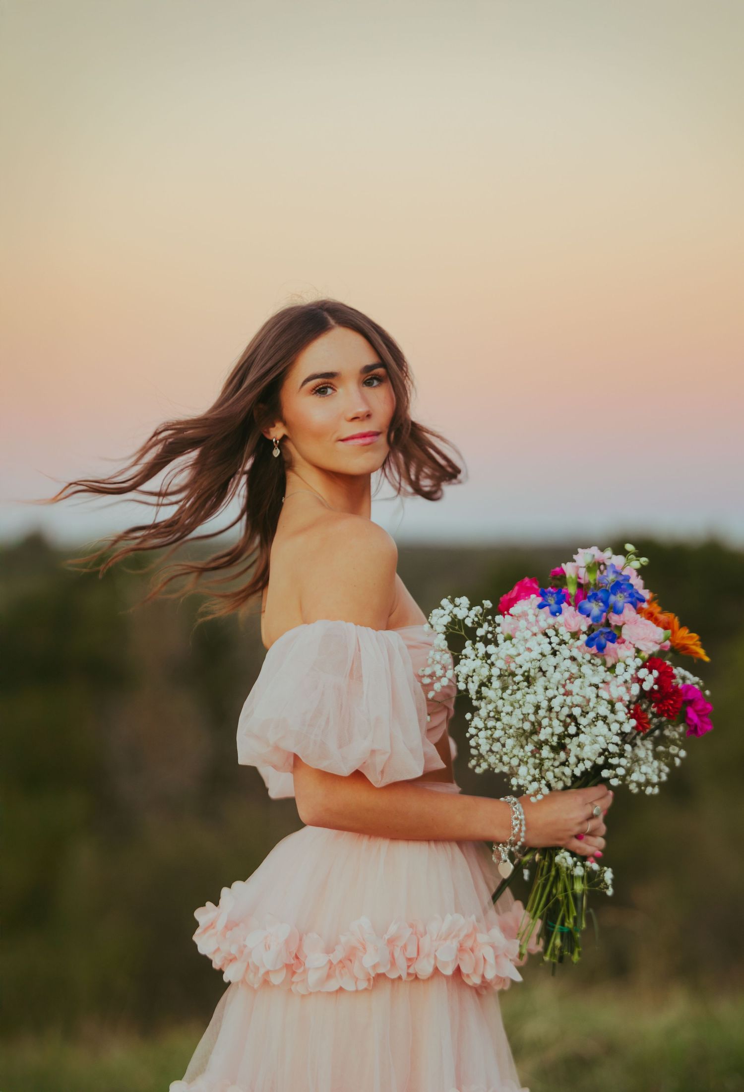 Brunette woman in pink off-shoulder dress holding colorful wildflower bouquet at sunset.