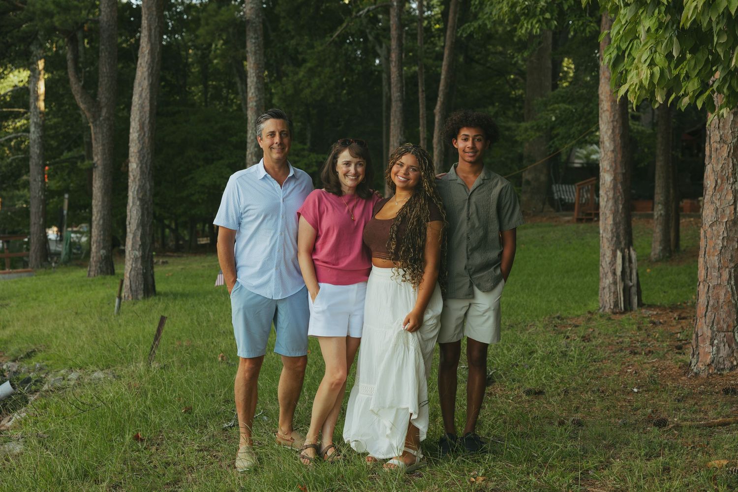 A family of four poses together smiling among tall pine trees on a sunny day outdoors. lake photos.