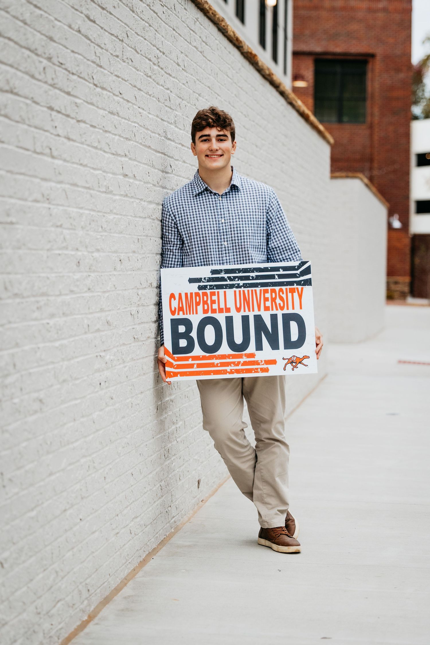 Smiling teen boy holding Campbell University Bound sign leaning against white brick wall.