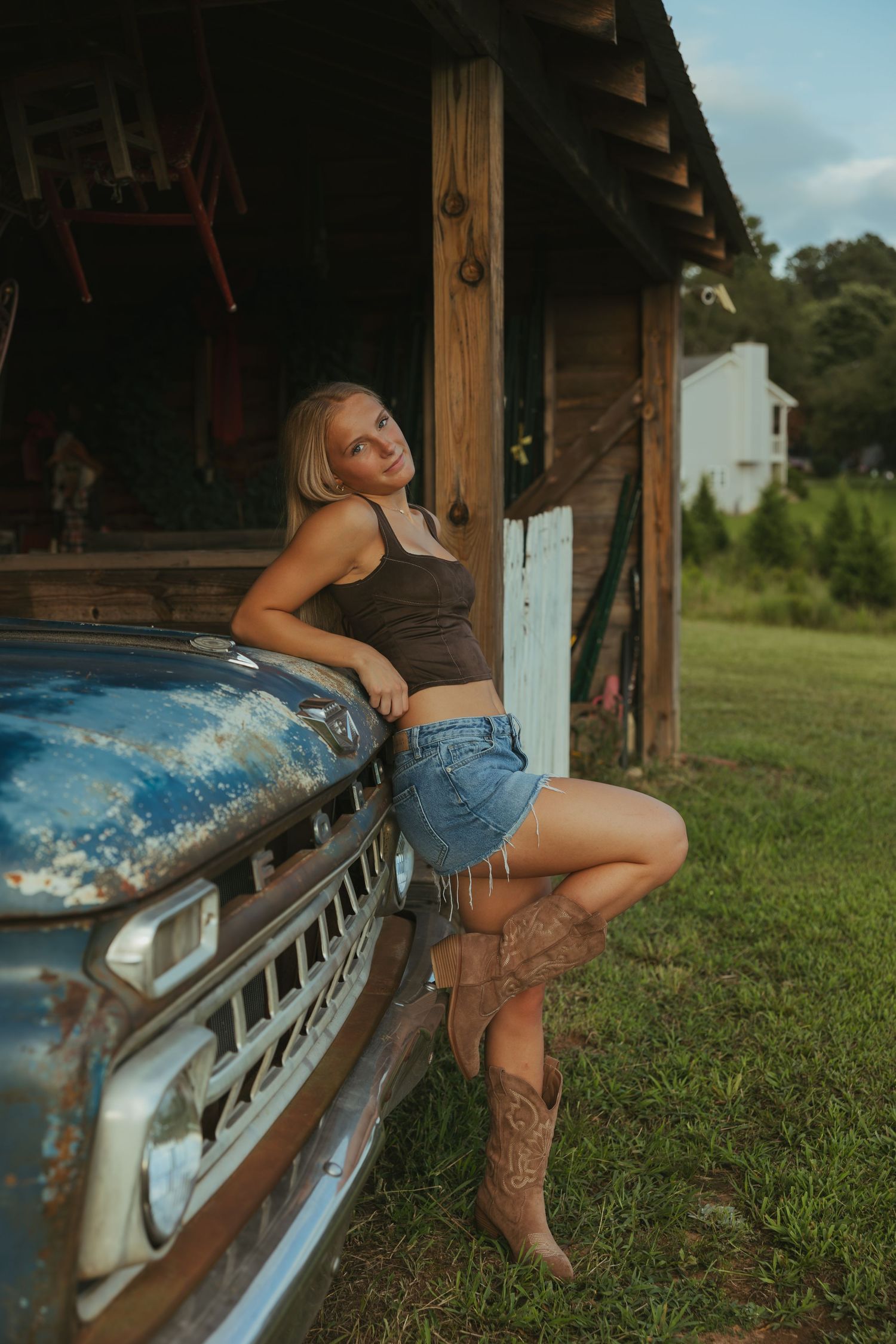Blonde woman in denim shorts and cowboy boots leaning on vintage truck by rustic barn.