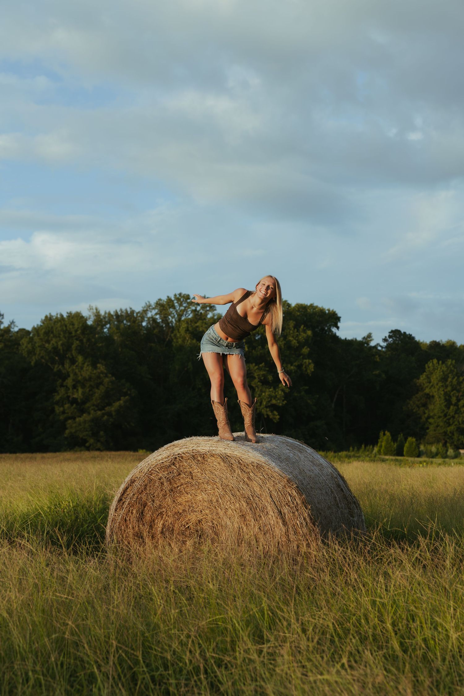Blonde woman posing on round hay bale in golden field at sunset with trees behind.