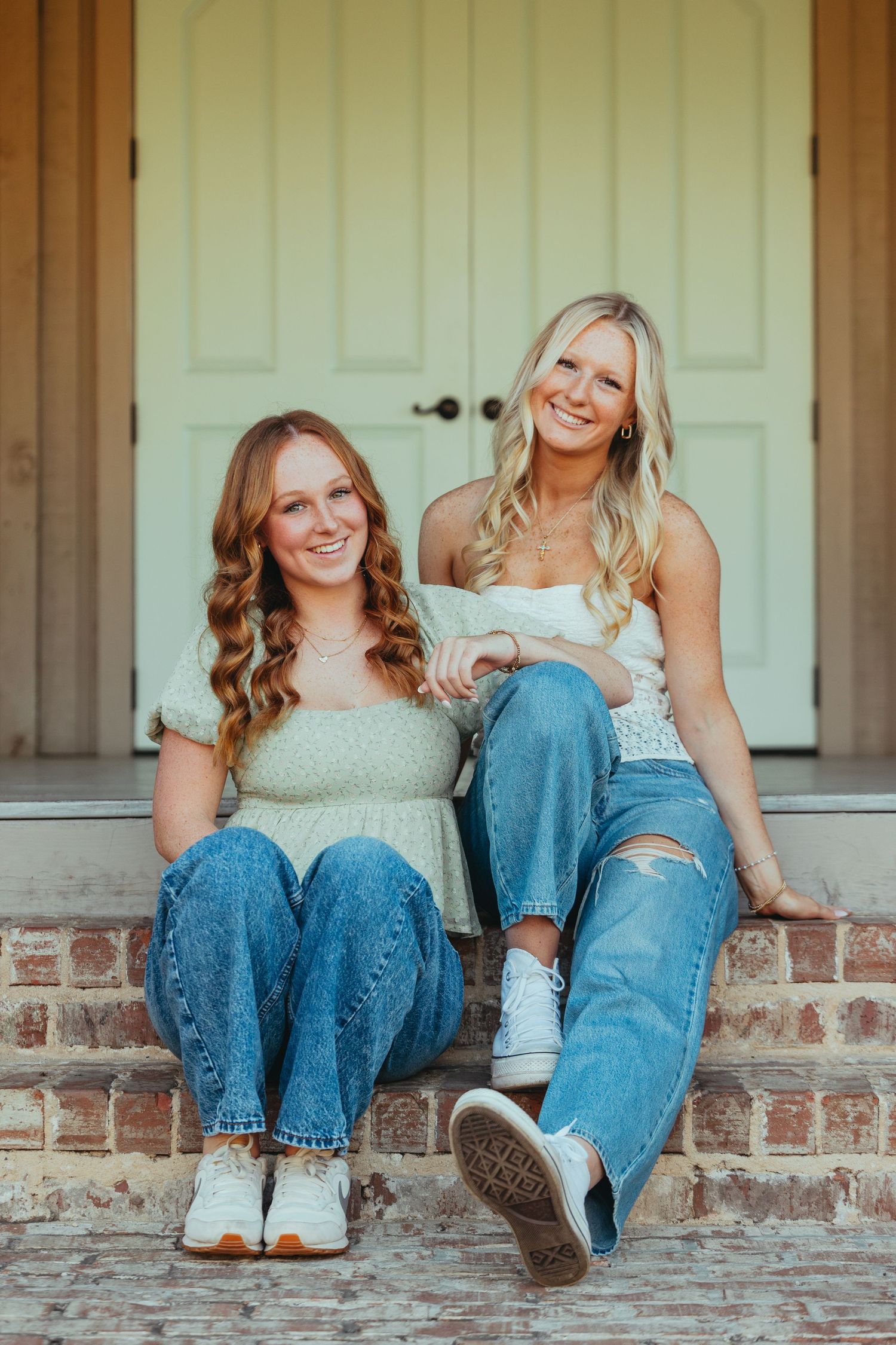 Two smiling graduates in jeans sit together on brick steps in front of a door.
