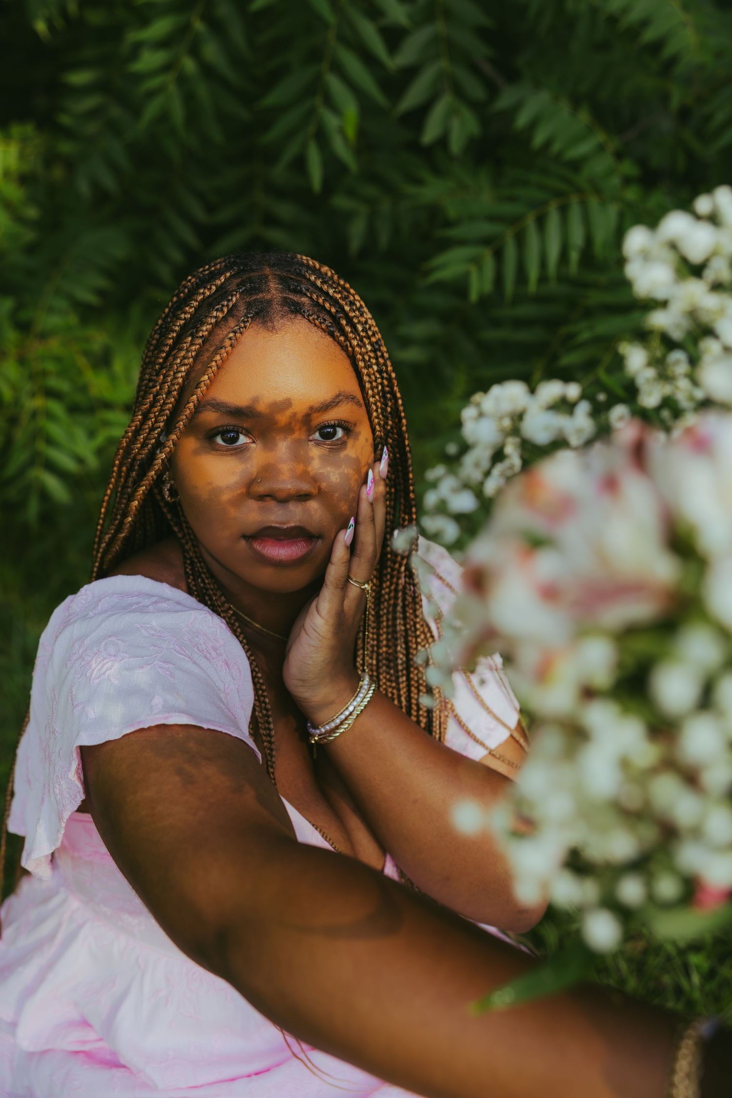 Young Black woman with braids in pink dress posing with flowers against lush green foliage.