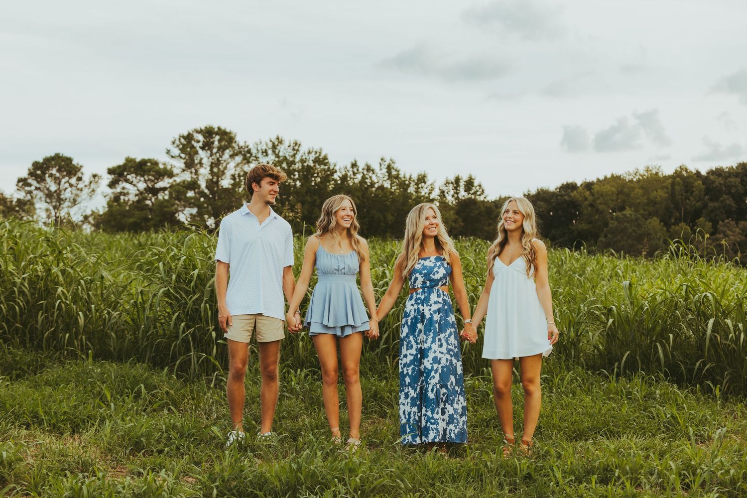Group of four young adults in blue outfits holding hands in a green field on a cloudy day.