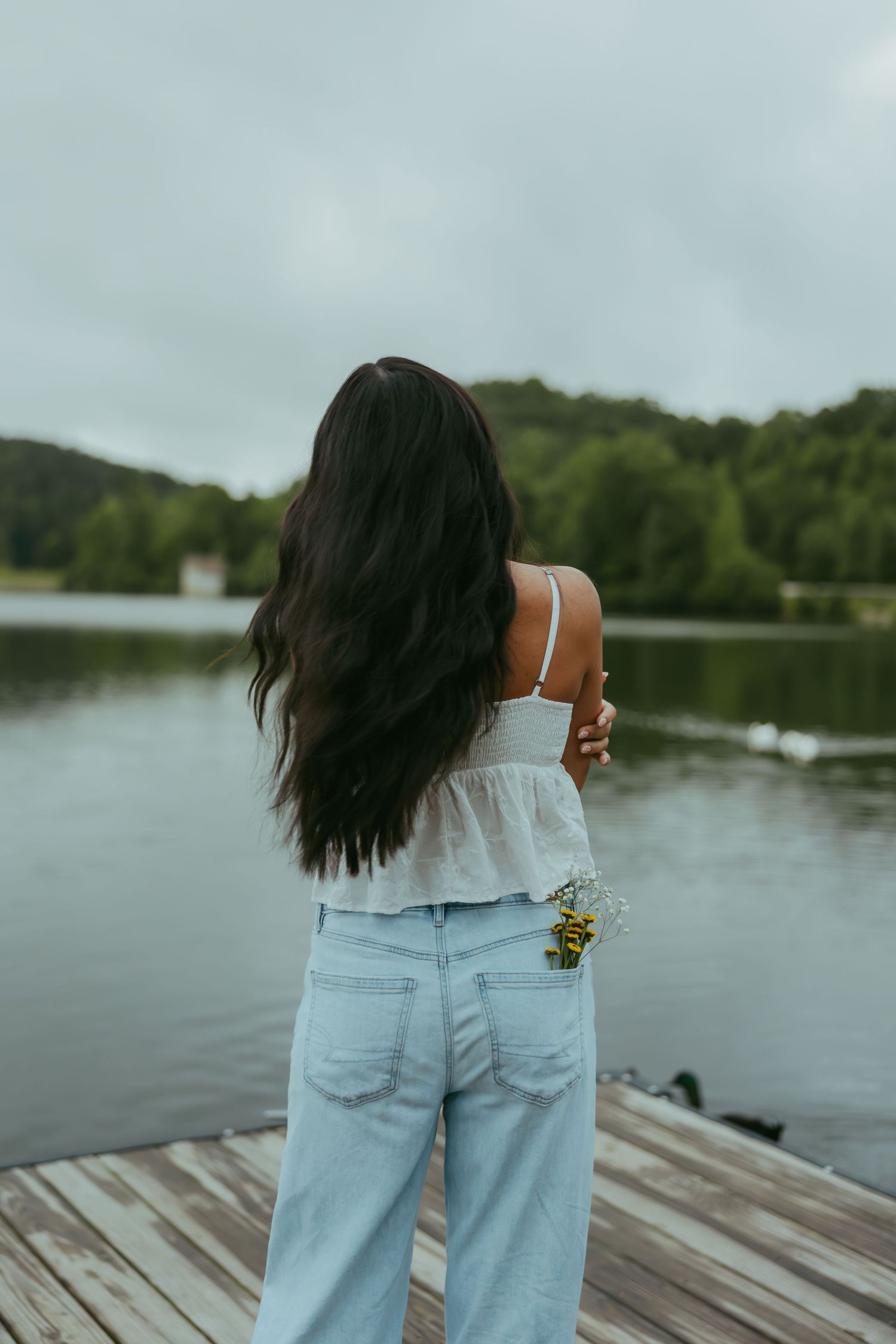 Young woman with long dark hair in white top and jeans holding flowers on wooden lake dock.