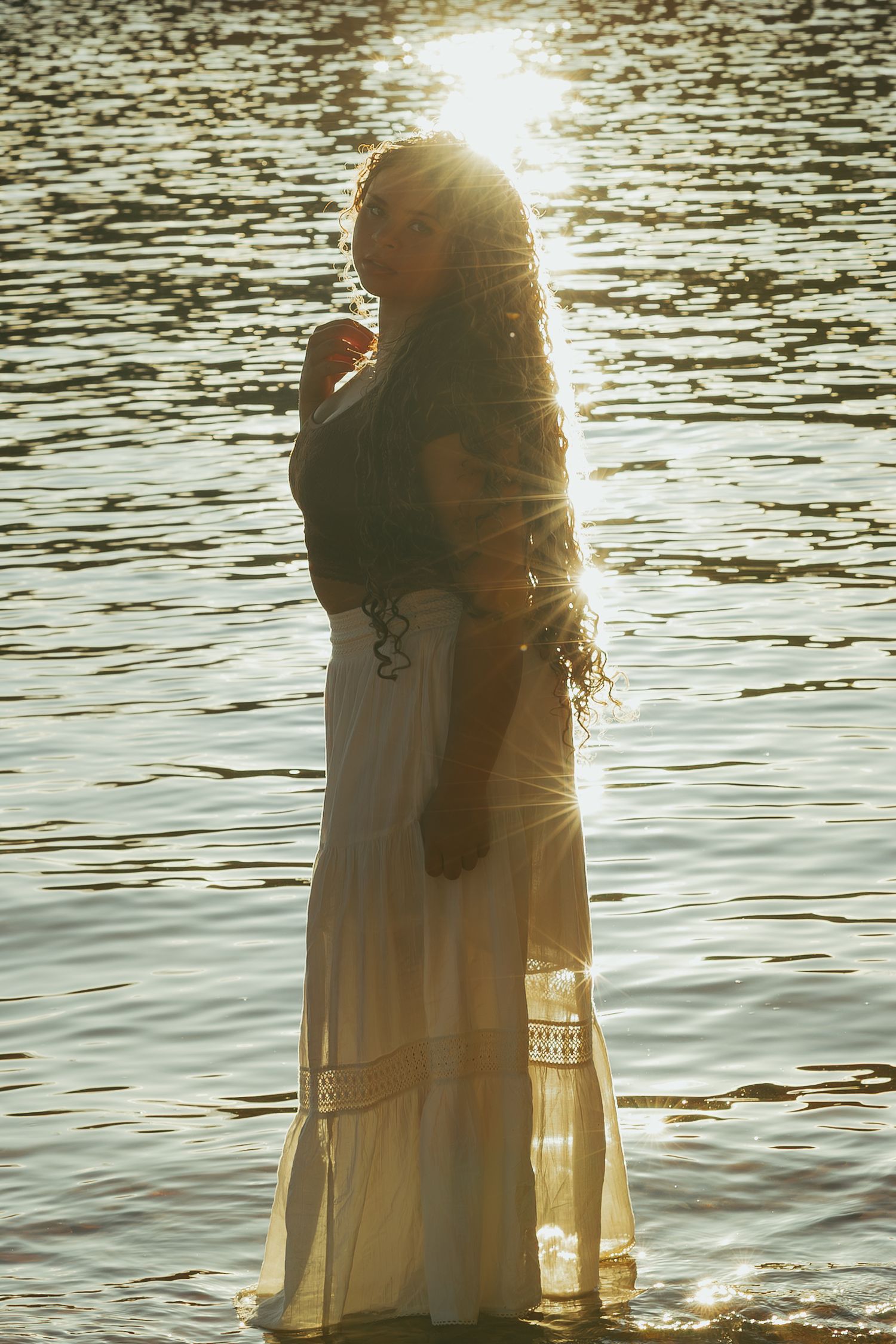 Woman in white maxi skirt standing in sunlit water with golden backlight reflection.