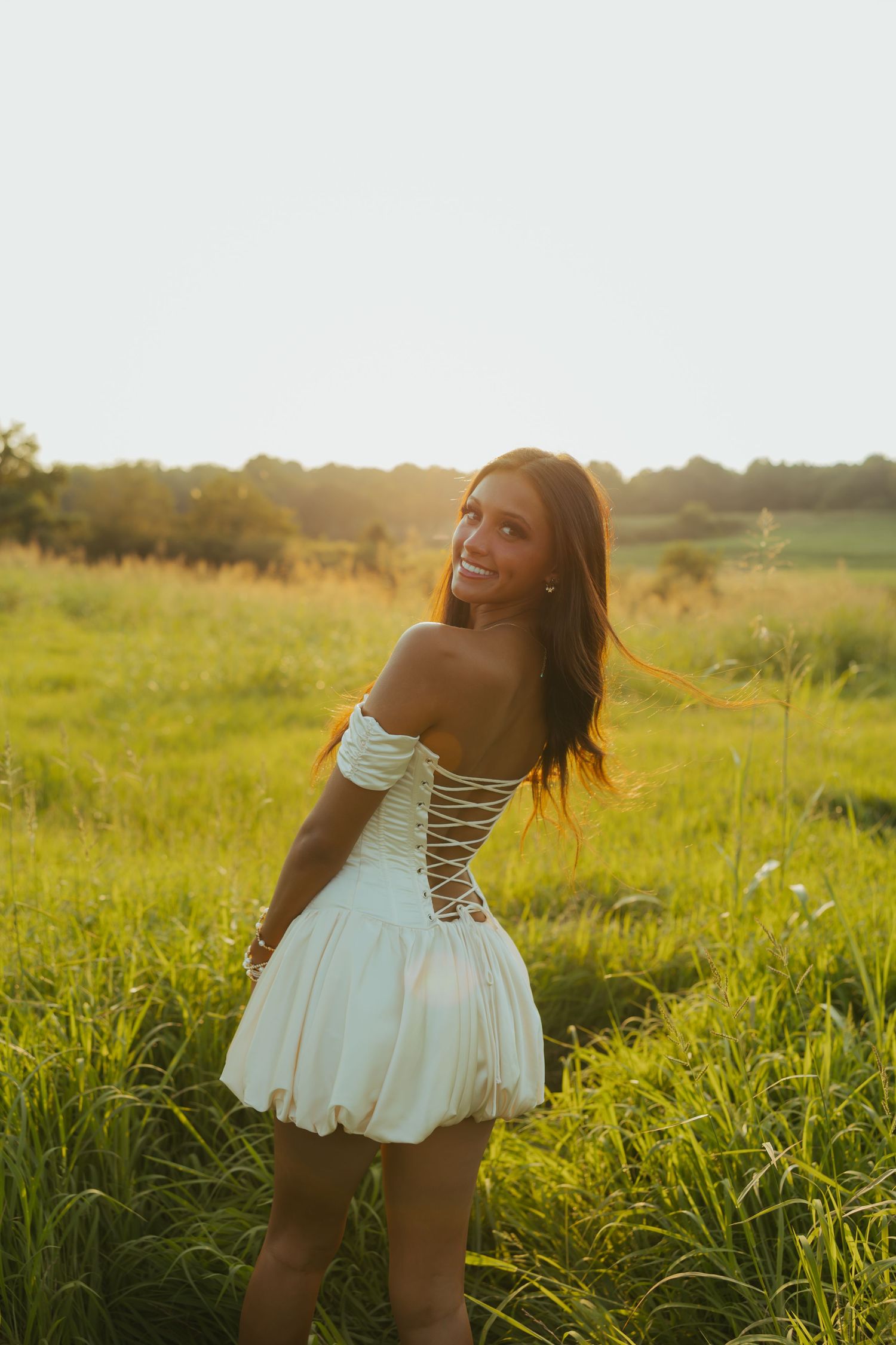 Young woman in white off-shoulder lace-up mini dress smiling in golden hour sunlit green field.