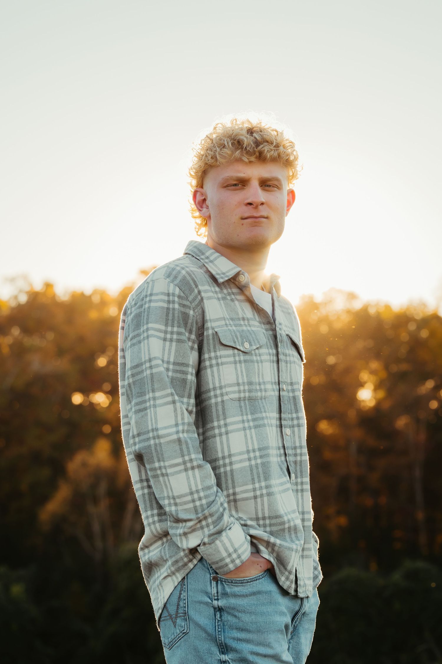 Young man with curly blonde hair in plaid shirt stands outdoors at golden hour sunset.
