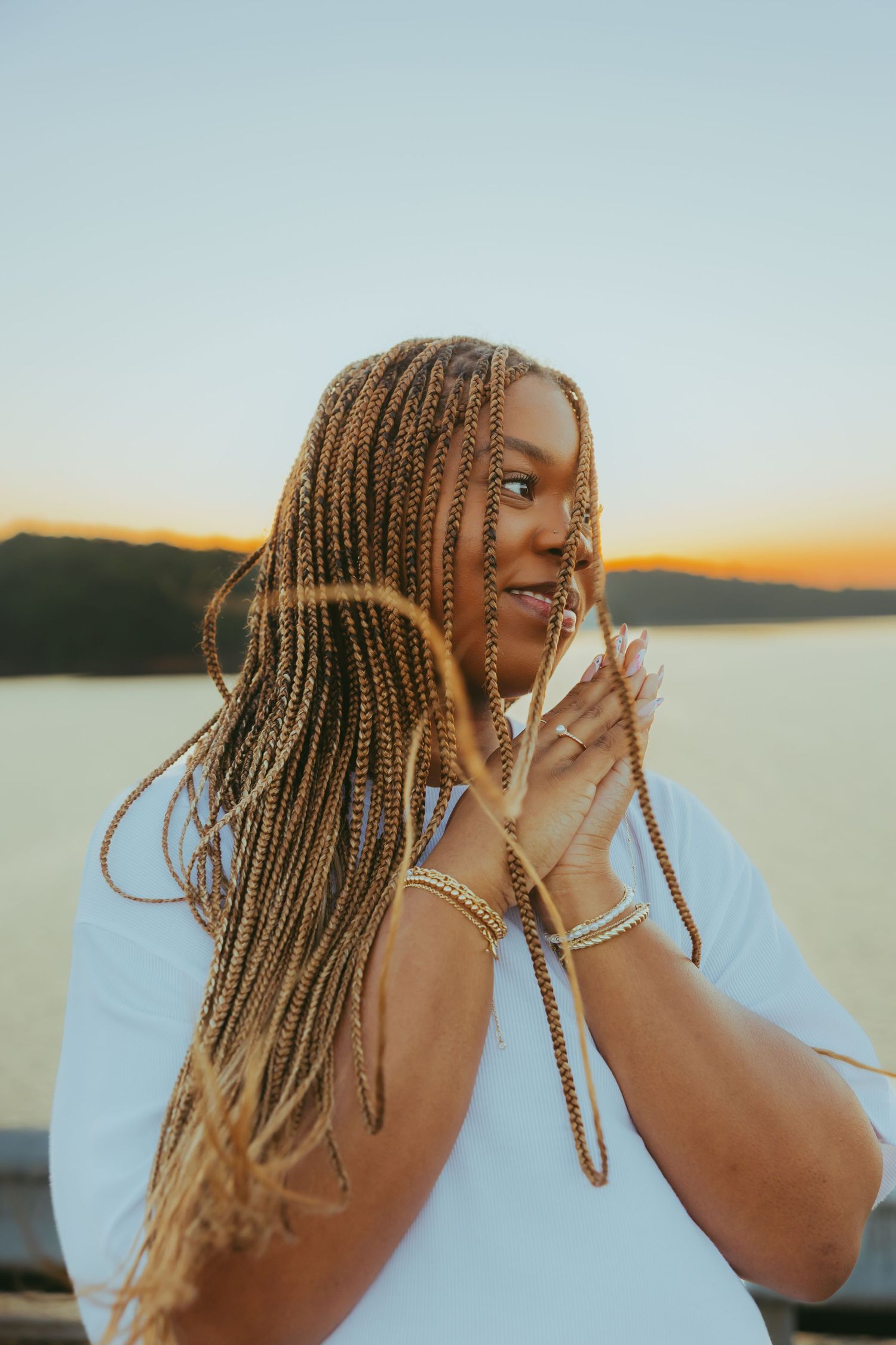 Young Black woman with long golden braids touching hair near waterfront during golden sunset.