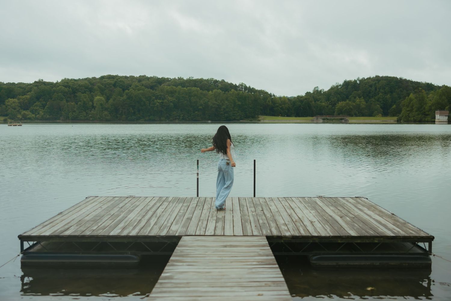 Woman in jeans standing on wooden dock overlooking calm lake surrounded by green forested hills.