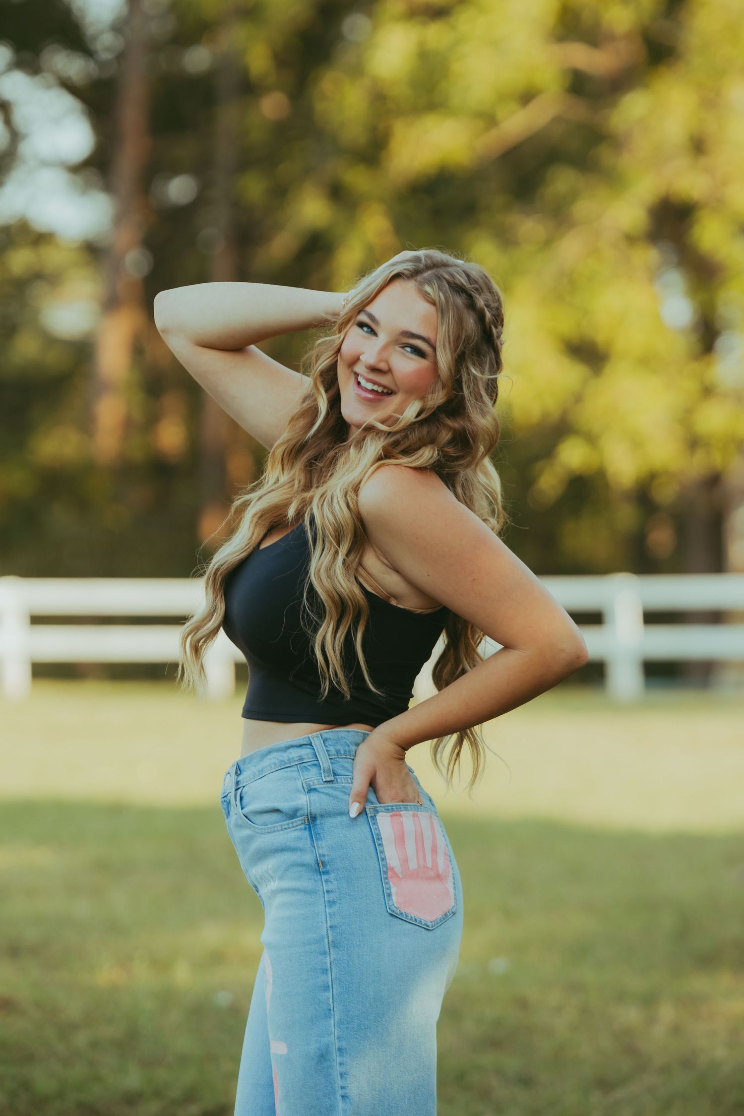 Smiling blonde woman in black crop top and jeans poses outdoors near a white fence at golden hour.