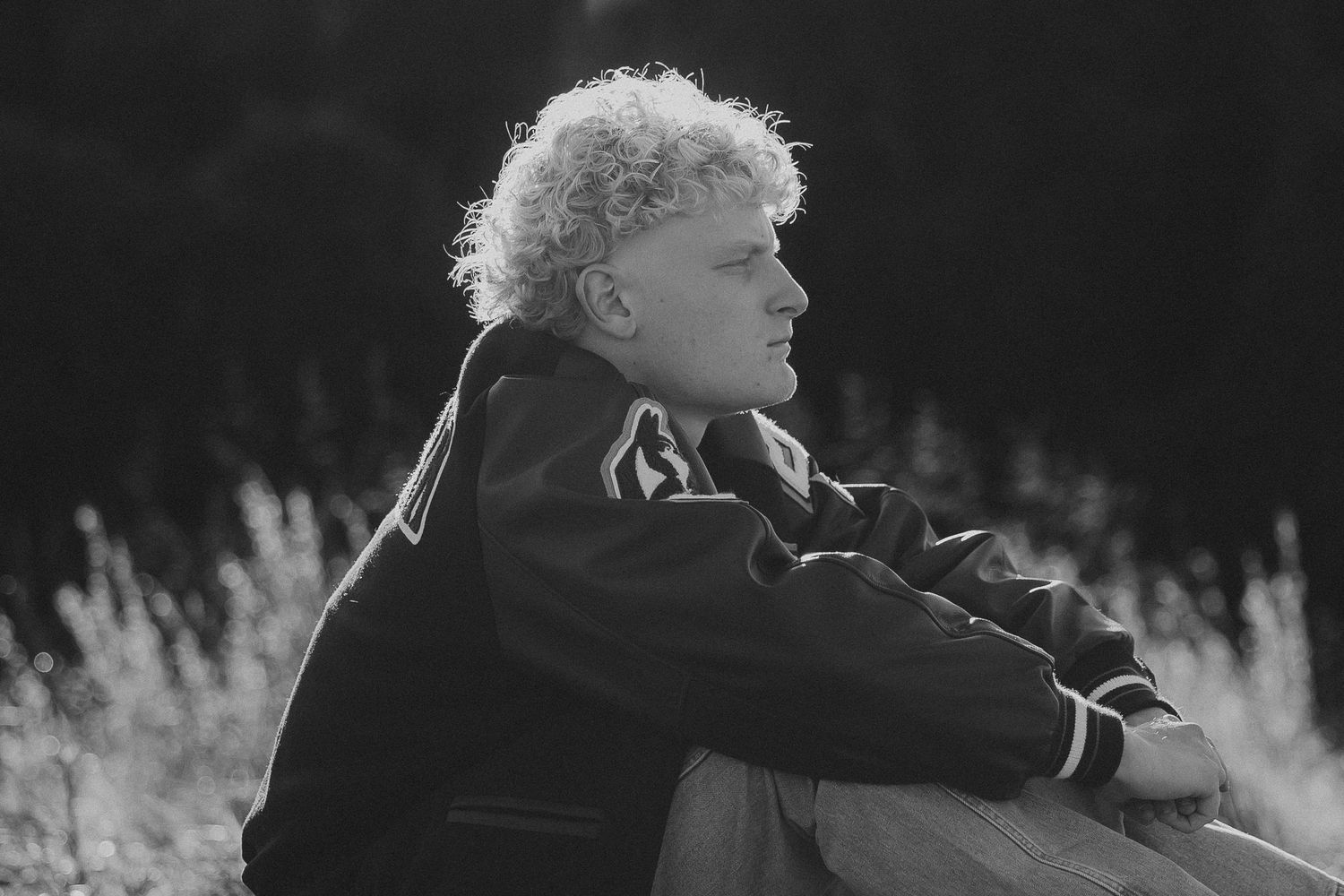 Black and white portrait of curly-haired young man sitting outdoors gazing into the distance.