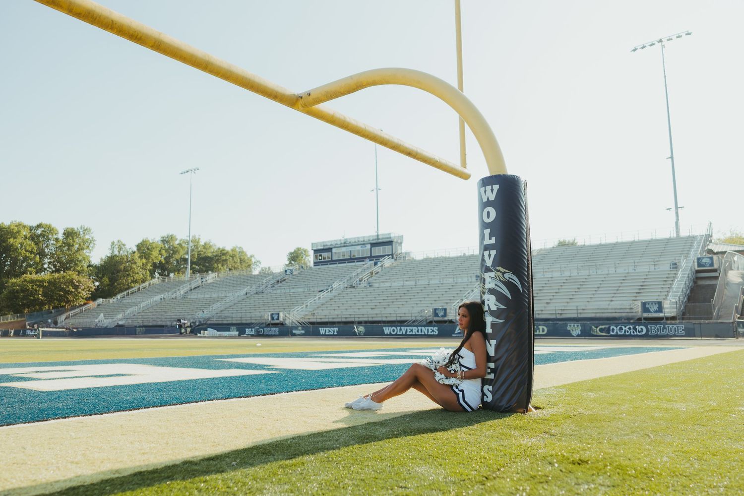 Female student posing beside football field goal post at Wolverines stadium on sunny day.